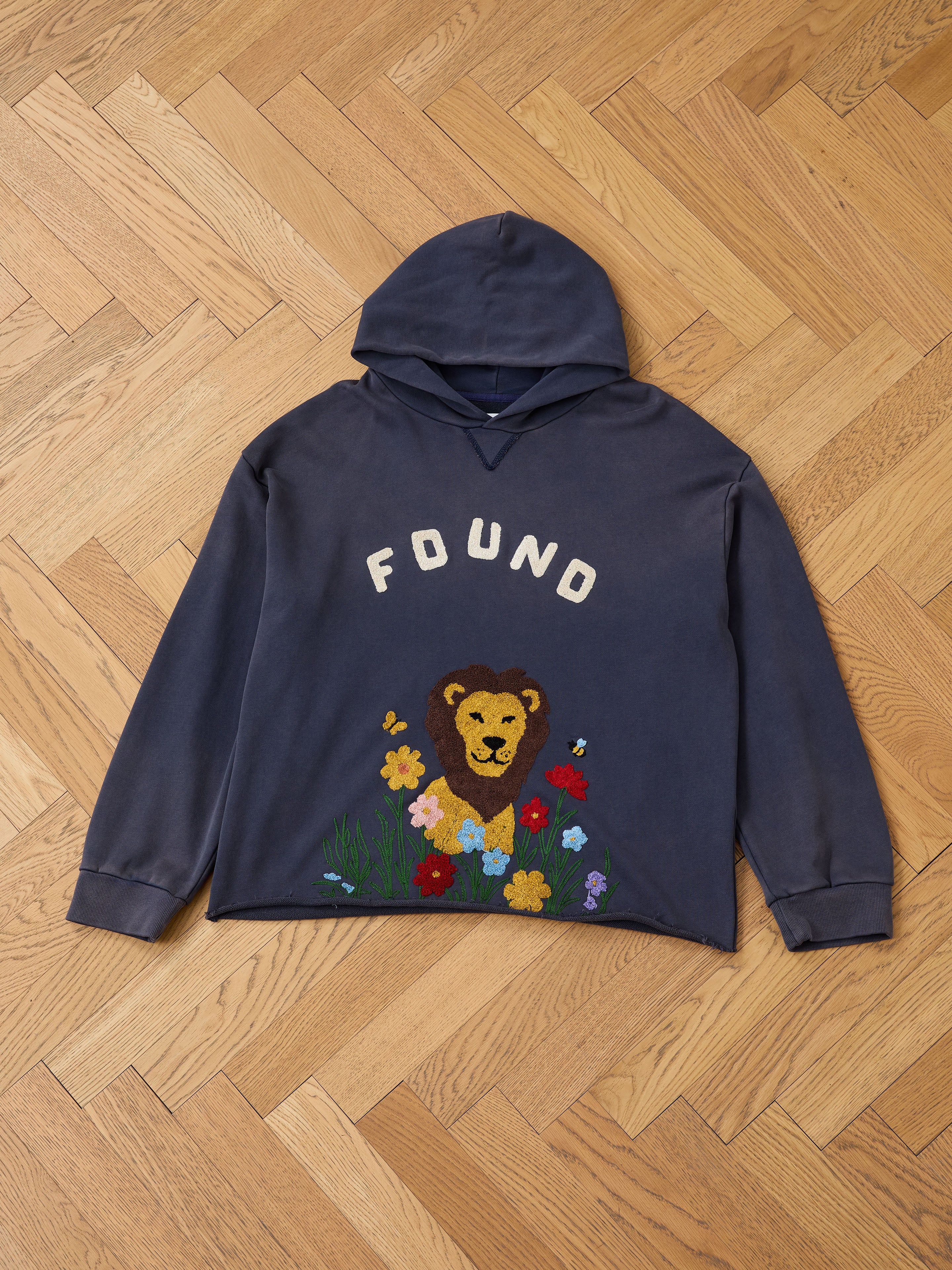 The FOUND Embroidered Wildlife Hoodie is a dark blue unisex relaxed fit, featuring "FOUND" above an embroidered lion's head and colorful flowers, shown flat on a wooden herringbone floor.