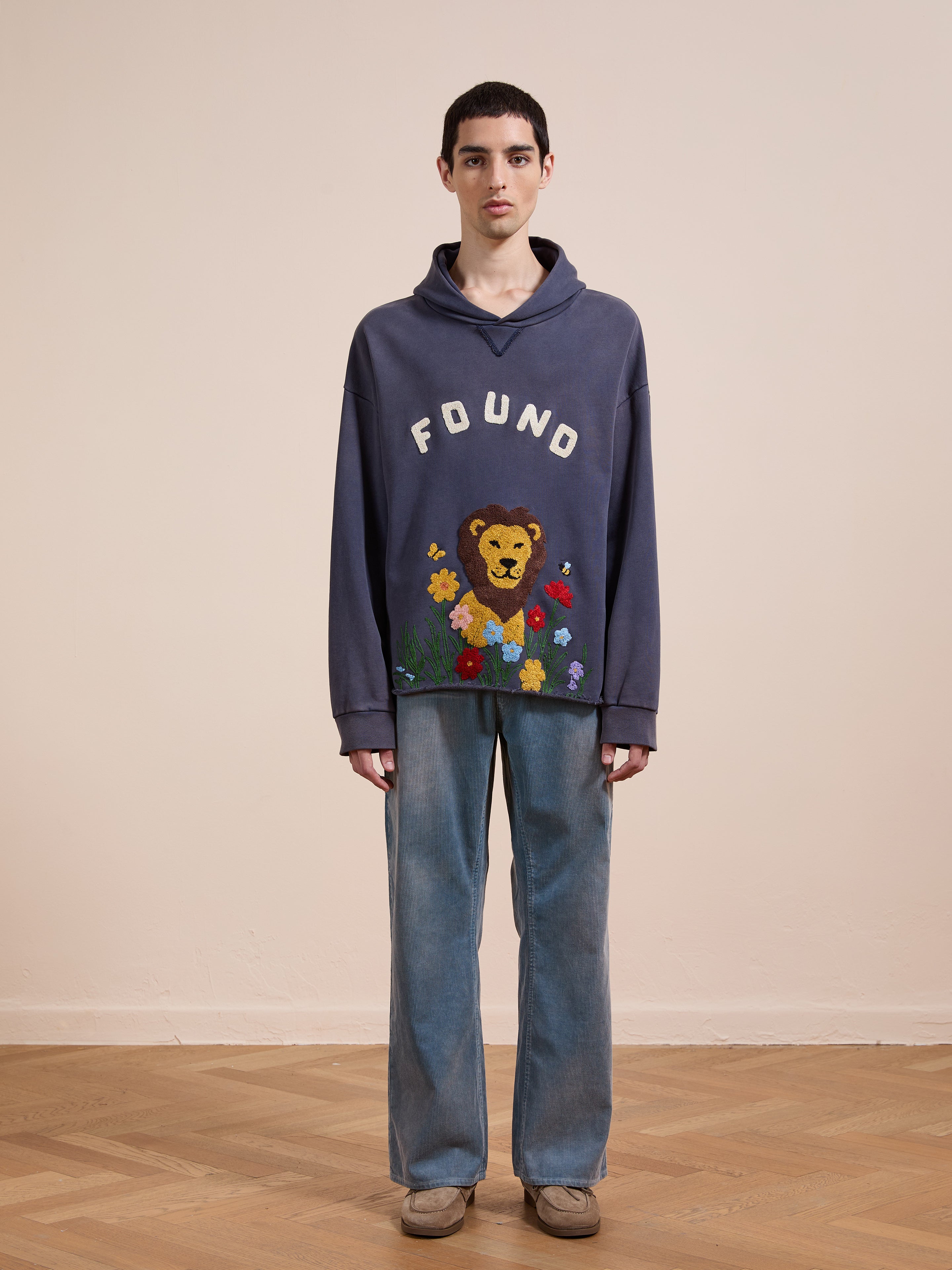 A person indoors wears the FOUND Embroidered Wildlife Hoodie, featuring a lion and flowers, paired with blue jeans and brown shoes, standing against a plain background.