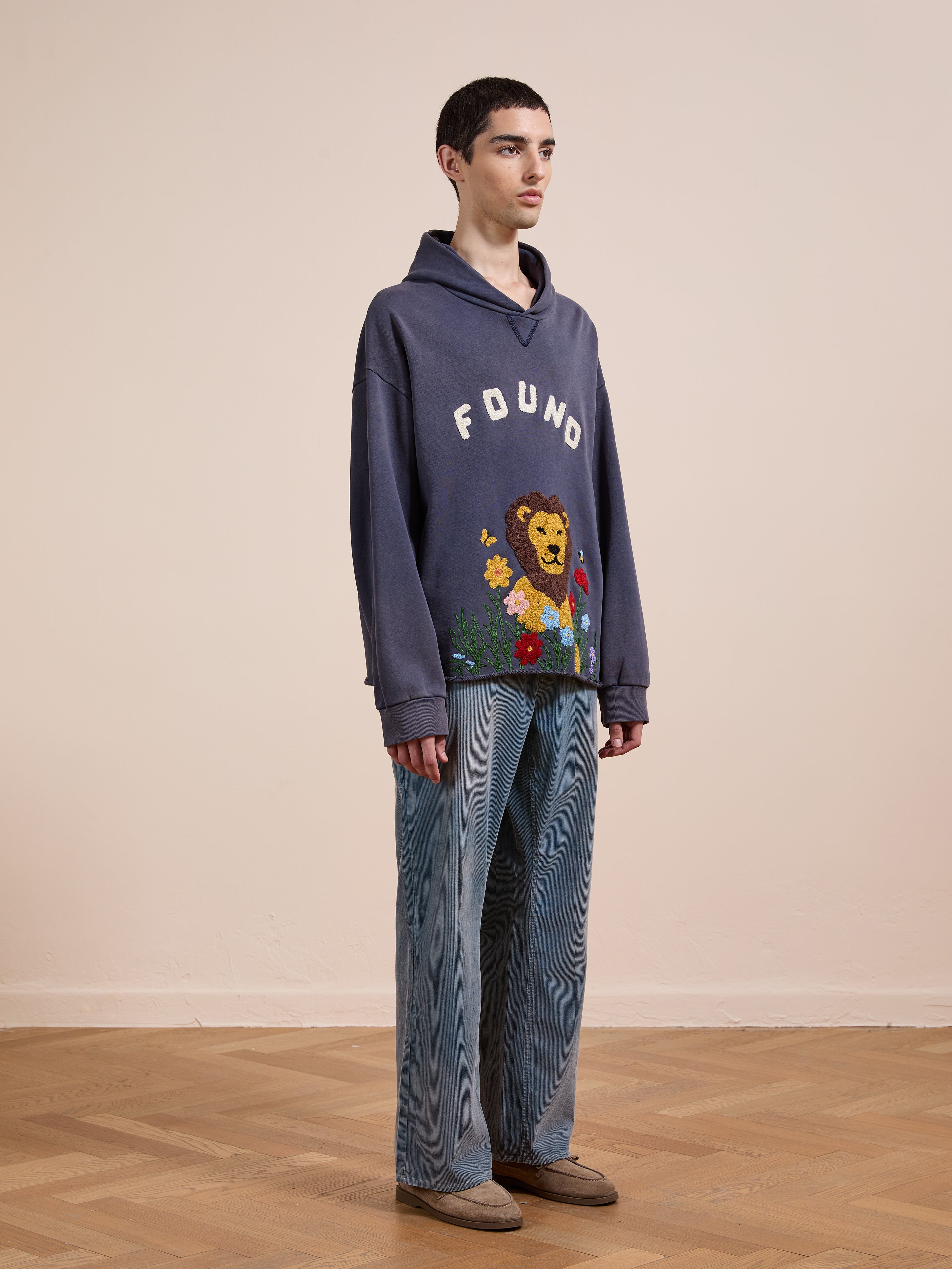 A person stands indoors on a wooden floor wearing the FOUND Embroidered Wildlife Hoodie with a lion graphic, paired with wide-leg jeans and brown shoes.