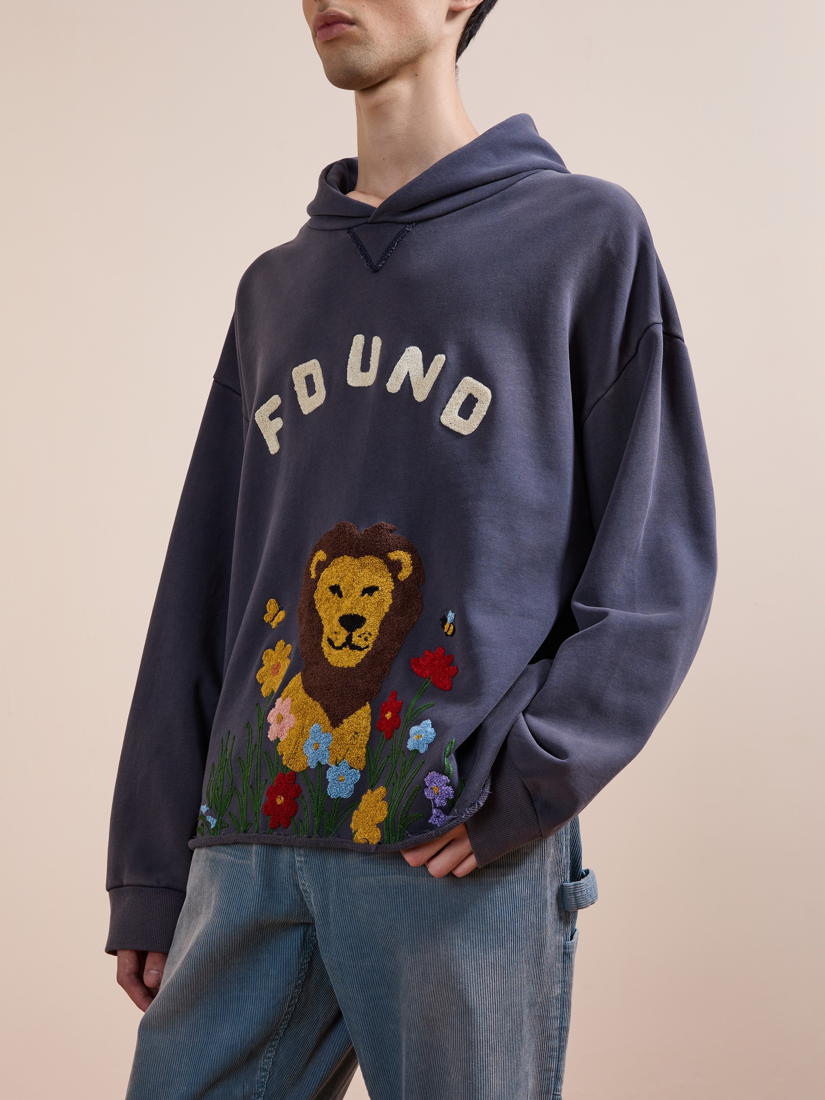 Someone wears the FOUND Embroidered Wildlife Hoodie featuring a lion face with flowers and "FOUND" text on the front, paired with light blue jeans.