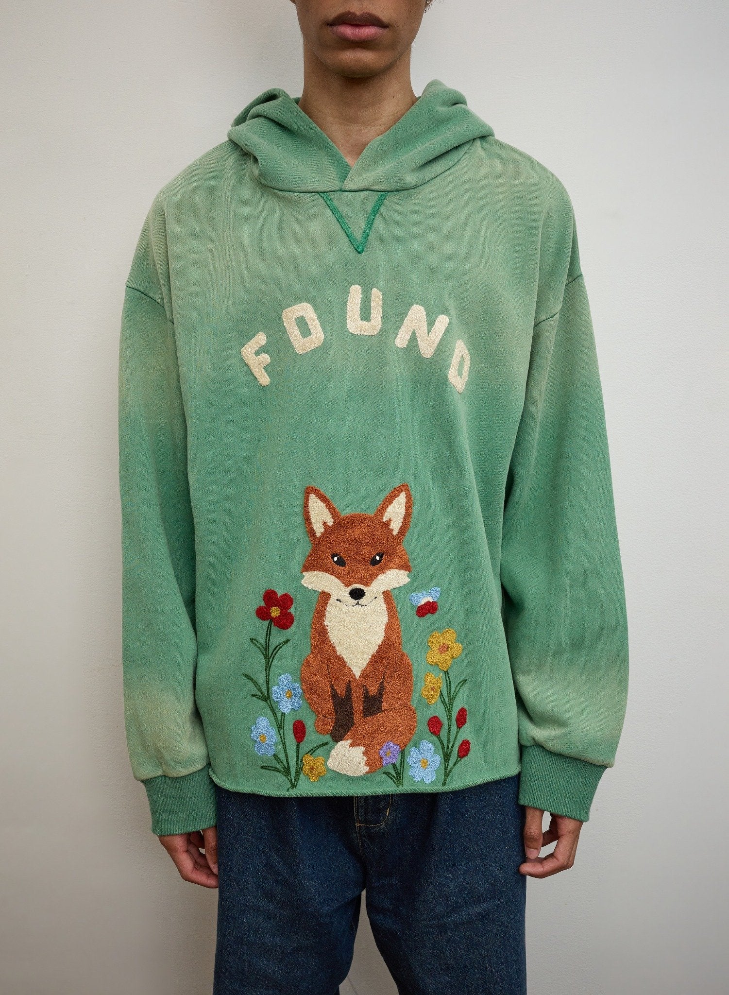 A person wears the FOUND Embroidered Wildlife Hoodie in green French-terry cotton, featuring a fox with flowers and "FOUND" embroidery, styled with blue jeans.