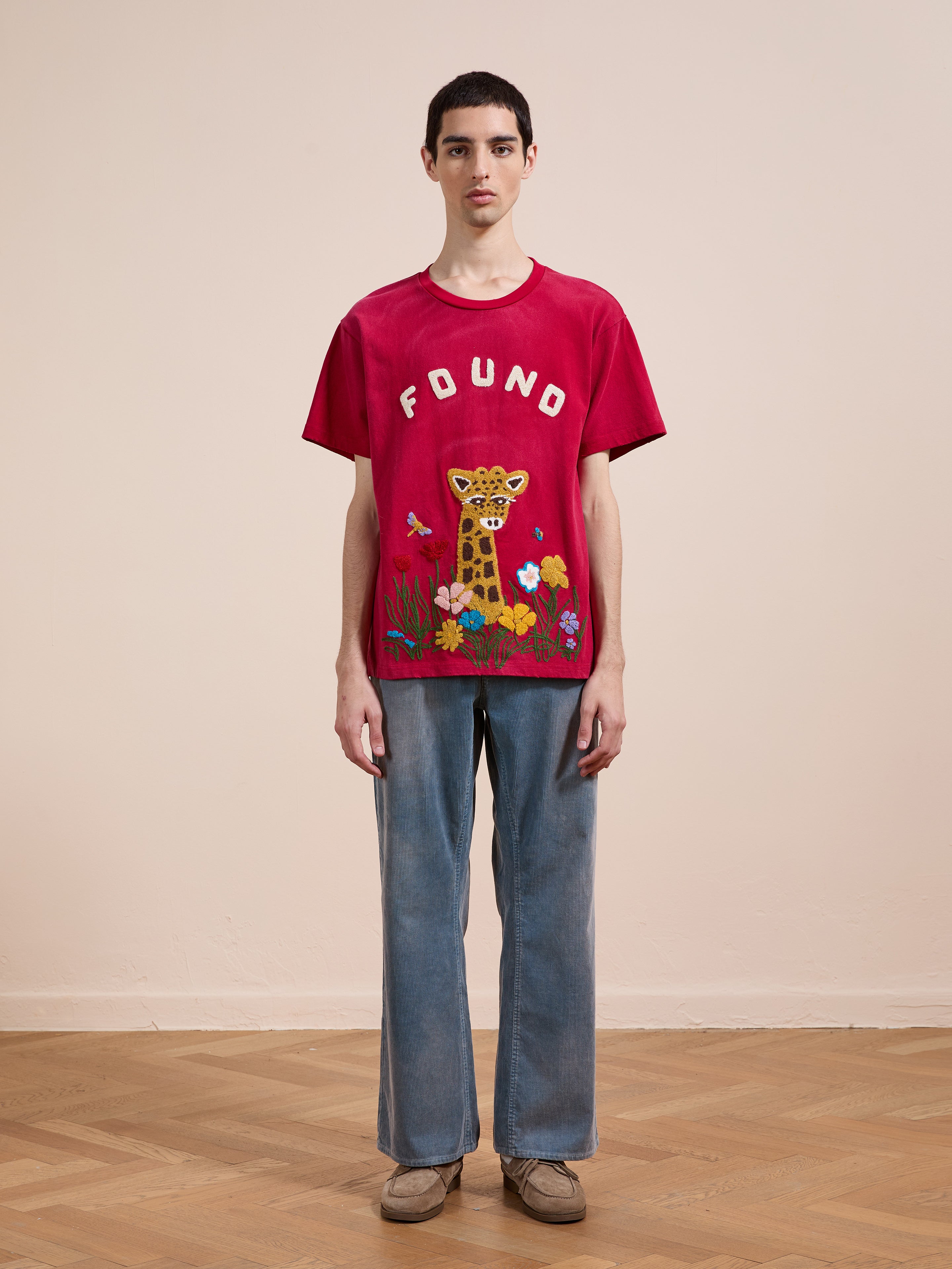 Indoors, a person wears the FOUND Embroidered Wildlife Tee, featuring a giraffe and flowers design, styled with blue jeans and beige shoes.