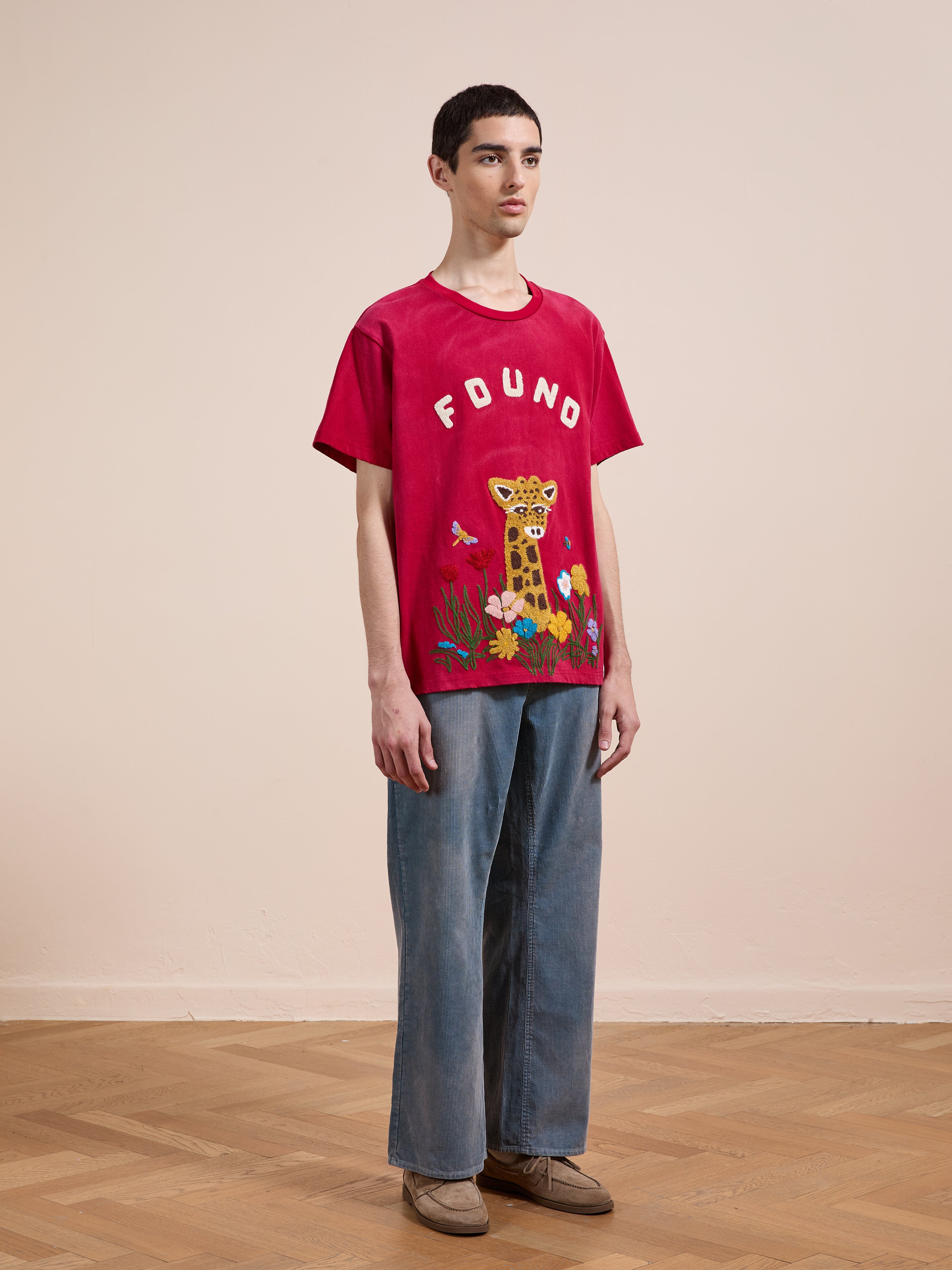 Indoors, a person wears the FOUND Embroidered Wildlife Tee with a giraffe design, styled with loose blue jeans and beige shoes for a relaxed, casual, unisex look.