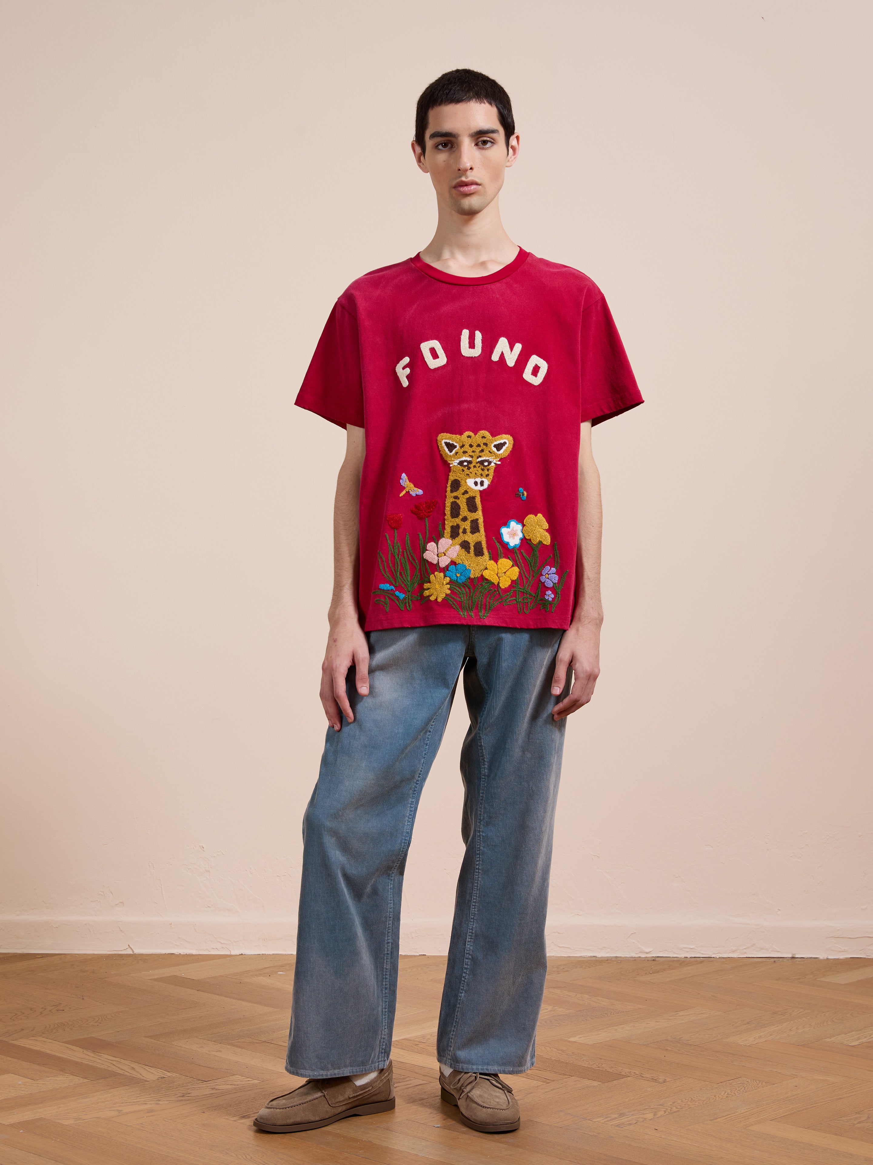 A person stands indoors against a light wall and wooden floor, wearing FOUND’s unisex Embroidered Wildlife Tee with a giraffe graphic, styled with blue jeans and brown shoes.