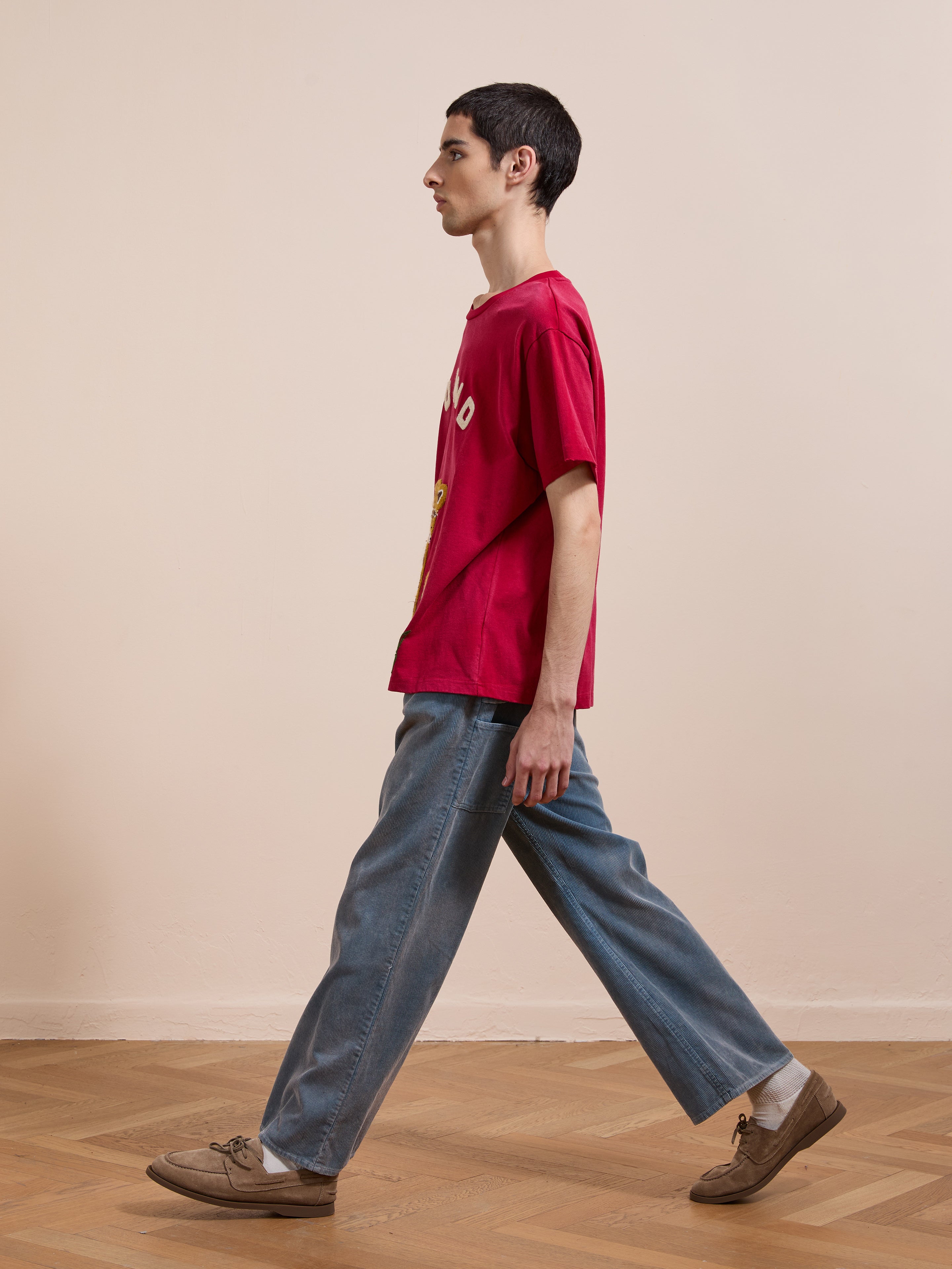 Wearing the FOUND Embroidered Wildlife Tee with blue jeans and brown shoes, a person walks indoors on a wooden floor against a plain beige wall.