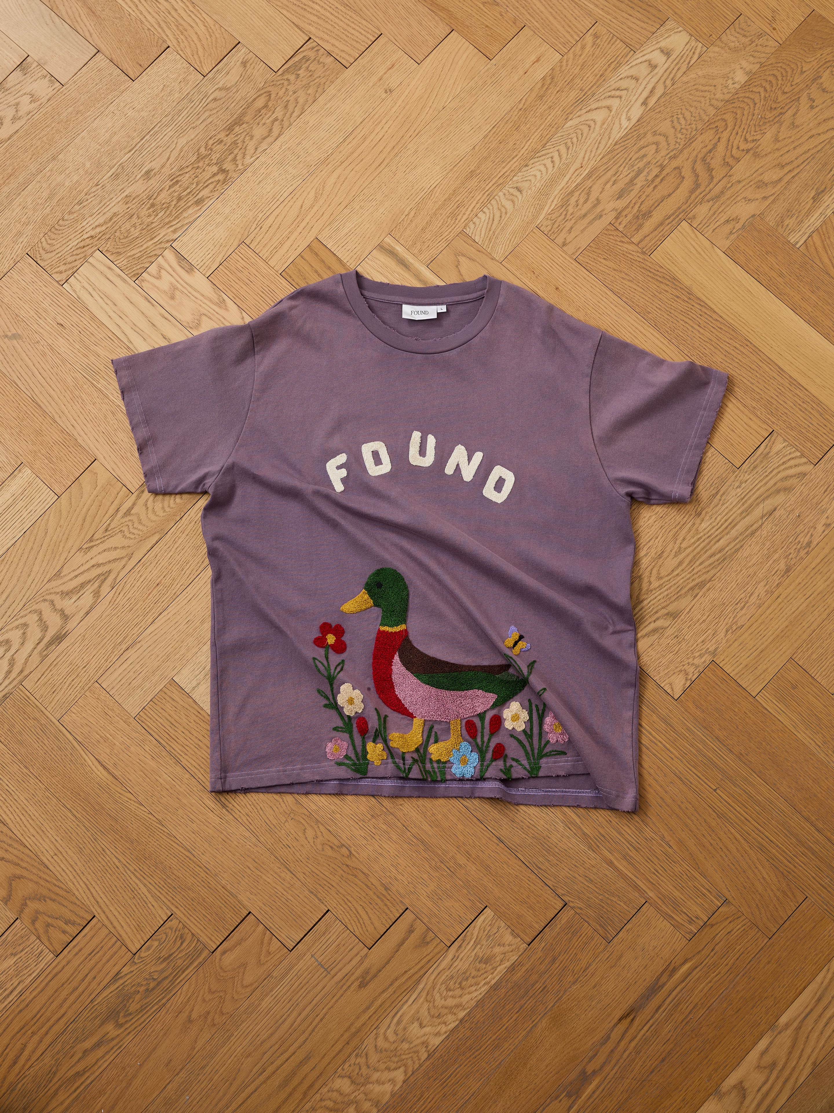 The FOUND Embroidered Wildlife Tee in purple features chenille embroidery with a duck and flowers design on the chest, displayed flat on a wooden herringbone floor.
