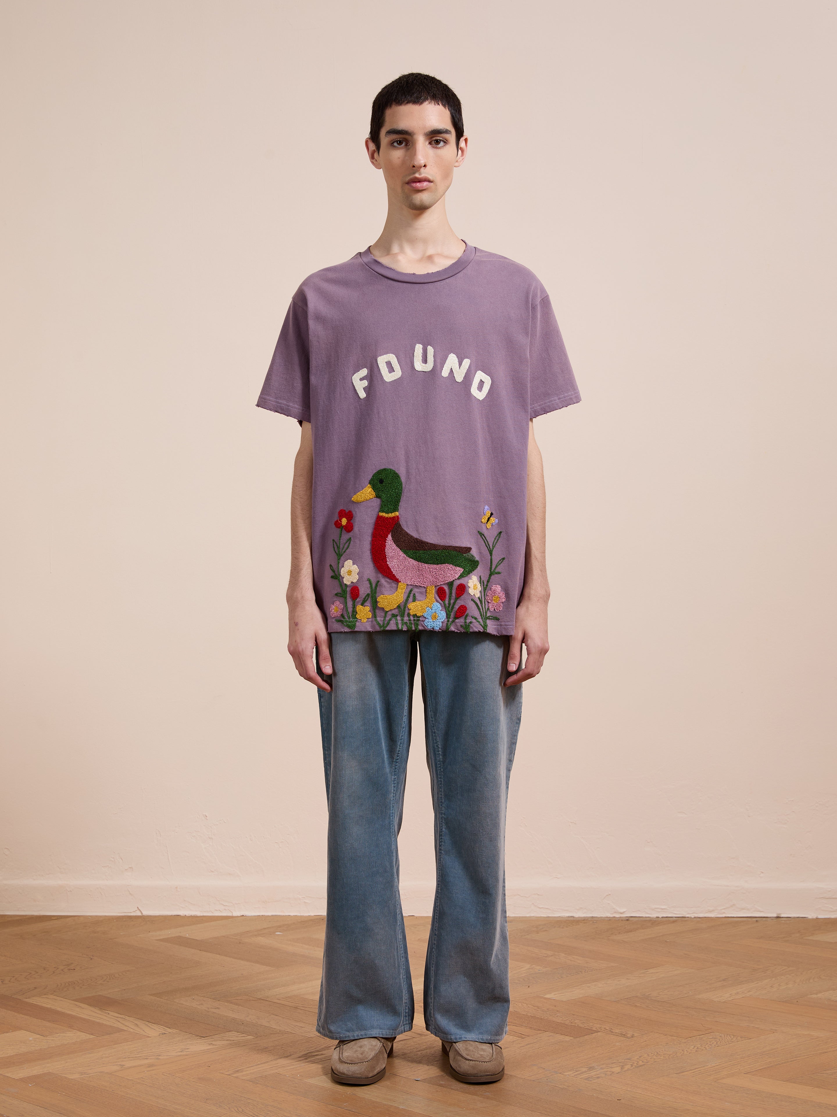 A person stands indoors wearing a purple FOUND Embroidered Wildlife Tee, blue jeans, and beige shoes against a plain background.