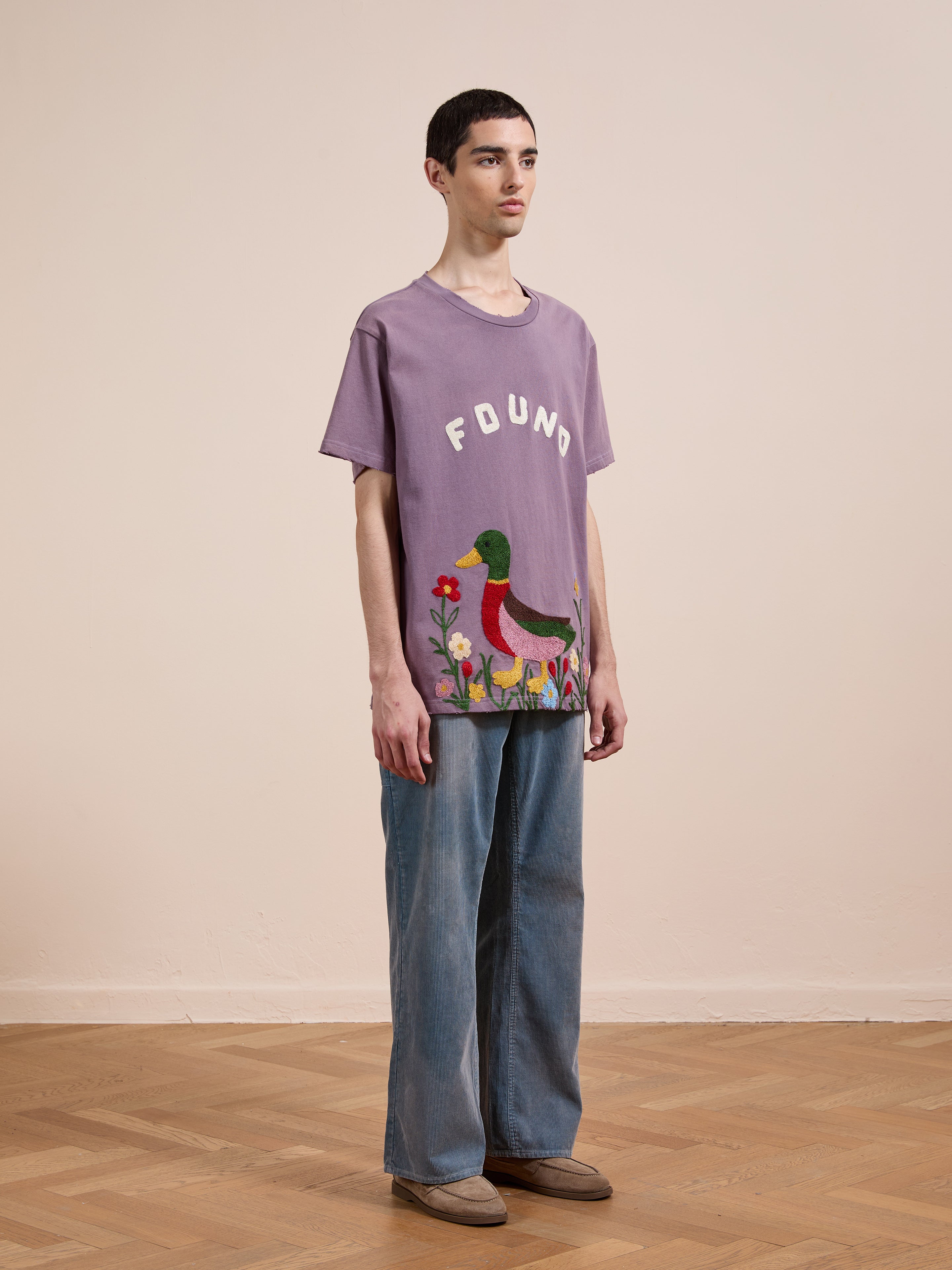 A person stands indoors wearing the FOUND Embroidered Wildlife Tee—a purple T-shirt featuring a duck and flowers—paired with wide-leg jeans and brown shoes.