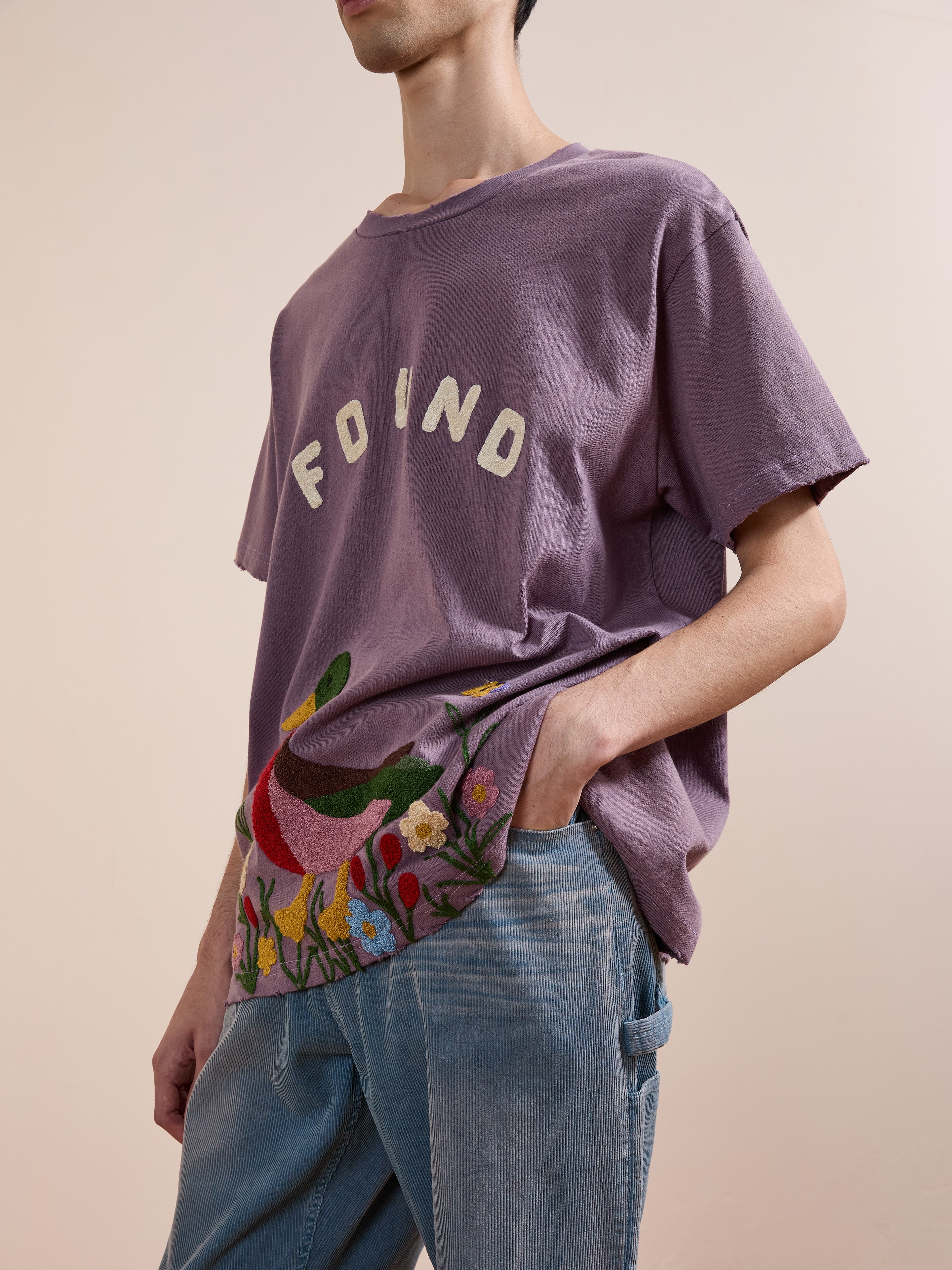 A person wearing the FOUND Embroidered Wildlife Tee, featuring purple chenille embroidery and floral details, paired with light blue jeans, stands against a beige background.