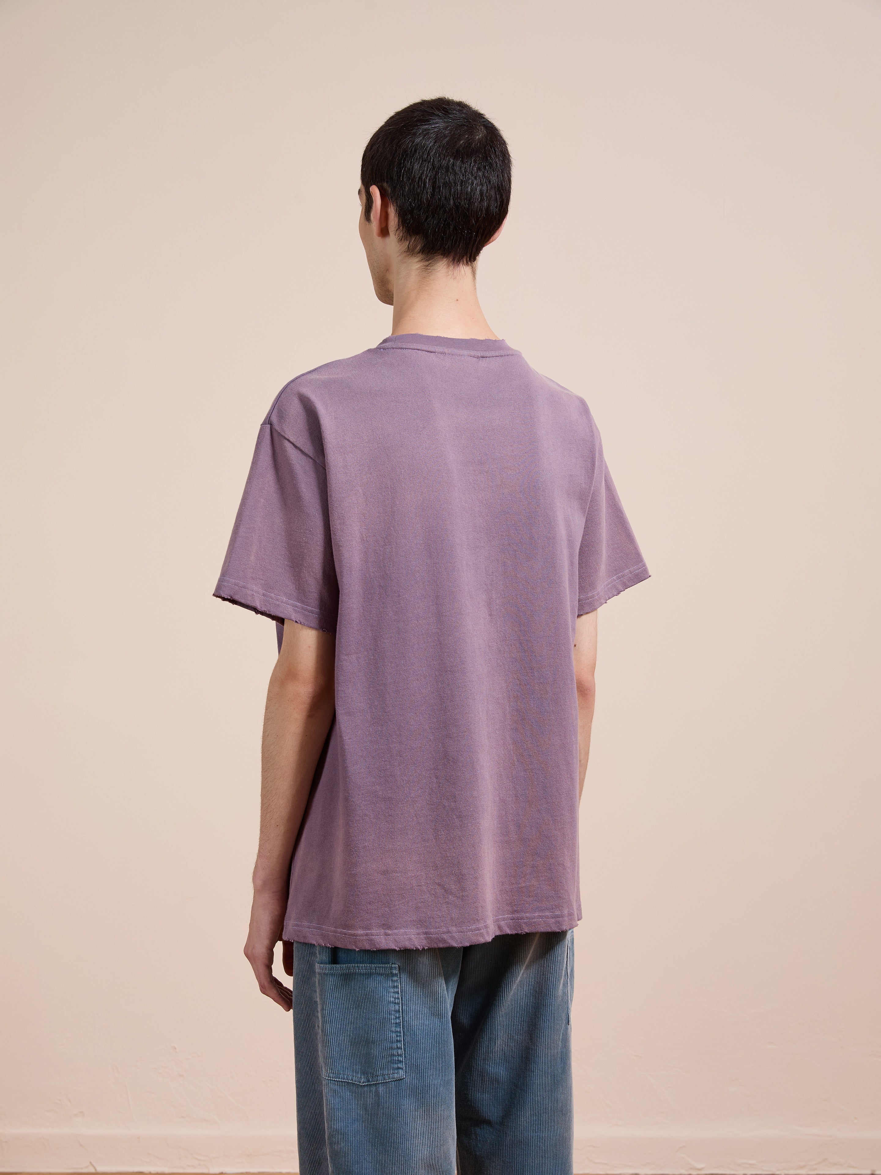 A person faces away from the camera, wearing a loose-fitting purple FOUND Embroidered Wildlife Tee and blue jeans against a plain beige background.