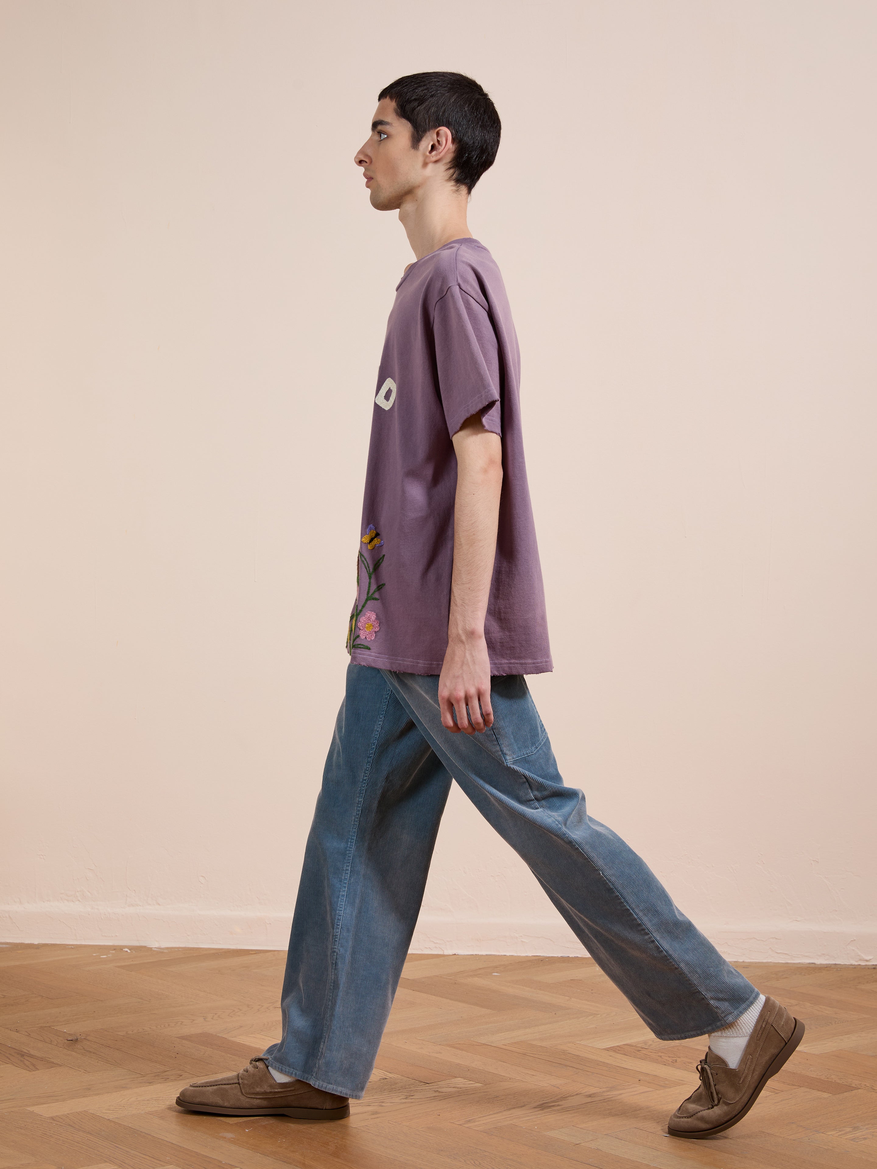 A person with short dark hair walks sideways indoors, wearing a loose FOUND Embroidered Wildlife Tee, wide blue jeans, and brown shoes on a wooden floor.