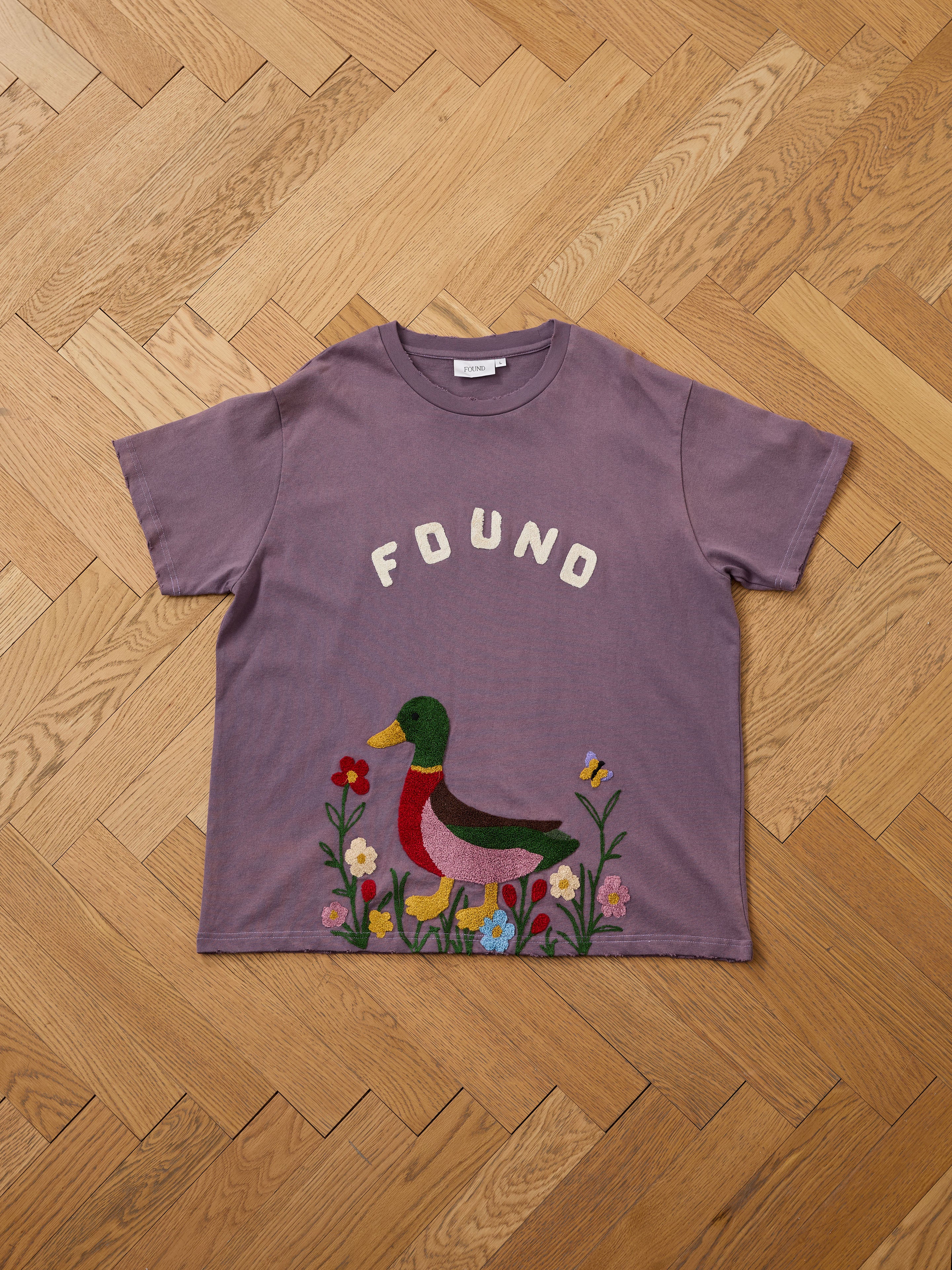 The FOUND Embroidered Wildlife Tee in purple features "FOUND" above a chenille duck, flowers, and a butterfly, shown laid flat on a wooden floor.