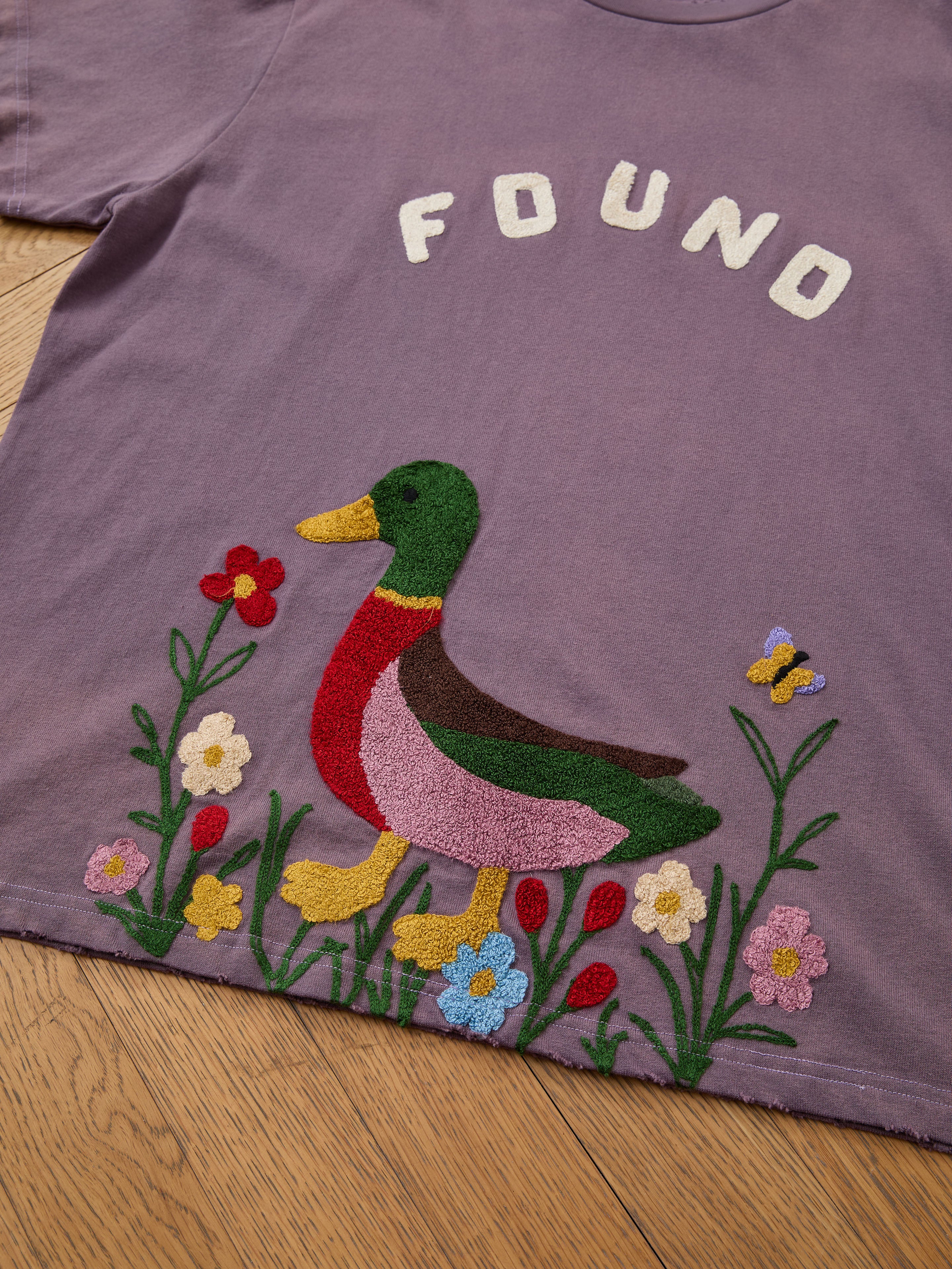The FOUND Embroidered Wildlife Tee features colorful chenille embroidery with a duck, flowers, and butterfly—perfect for nature lovers. Displayed on a wooden floor.