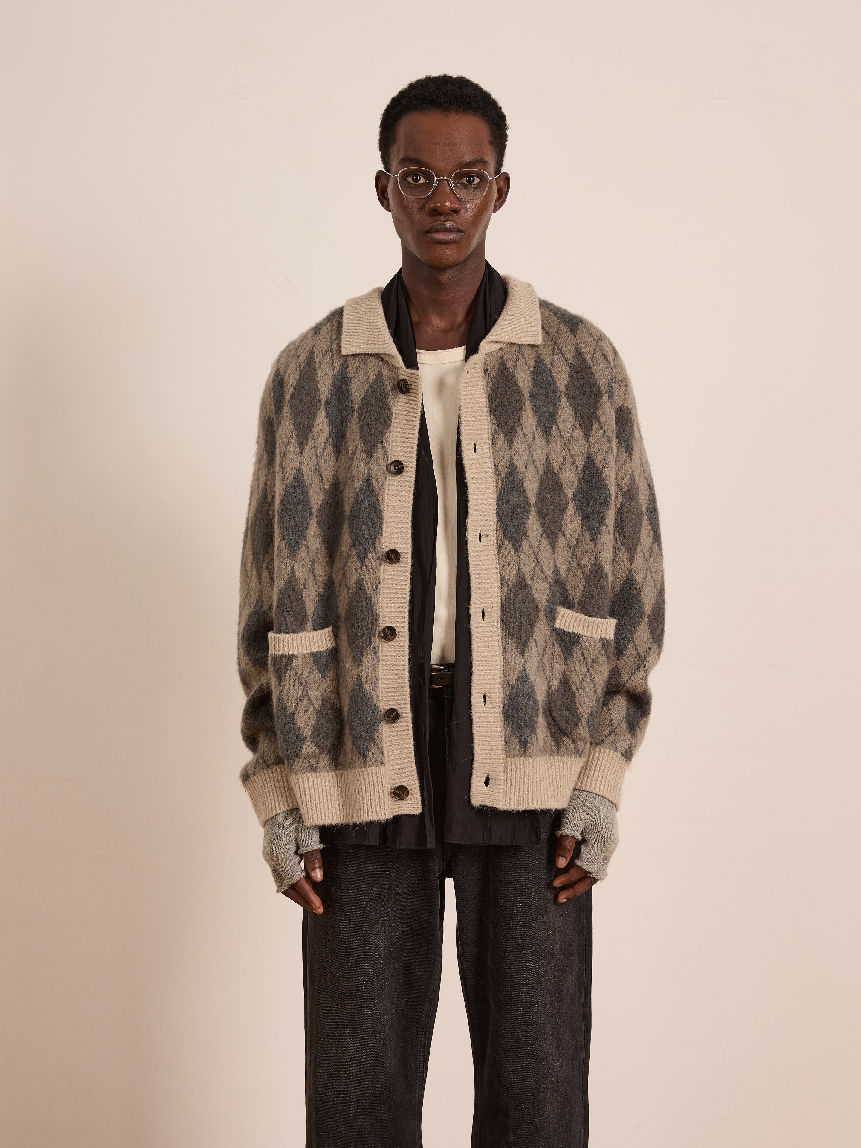 A person stands against a plain background wearing glasses, fingerless gloves, dark pants, a white shirt, and the FOUND Argyle Cardigan in a relaxed fit wool blend in beige and gray.