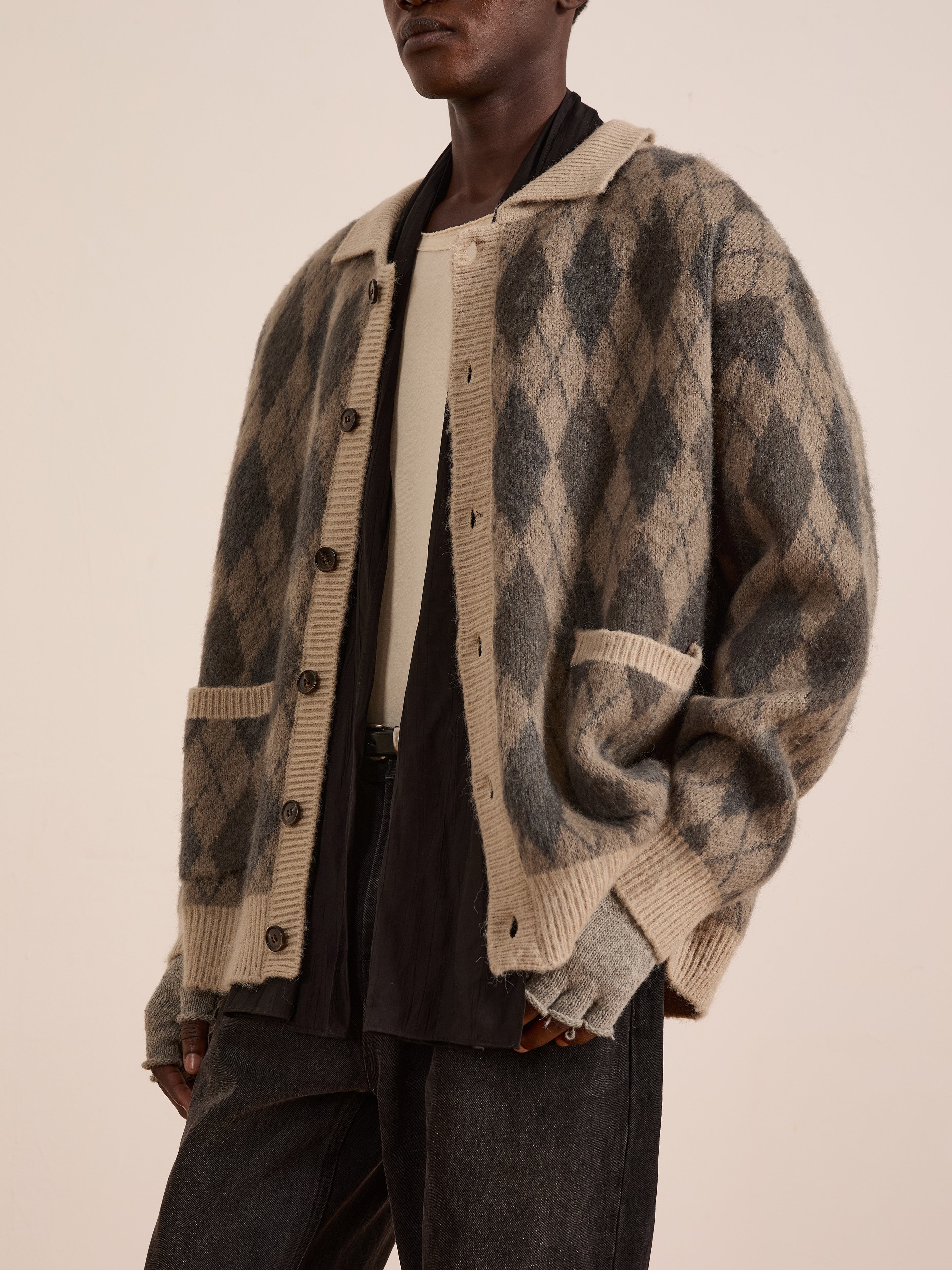 A person wears the FOUND Argyle Cardigan in beige and gray over a cream shirt and dark trousers, standing against a plain light background.