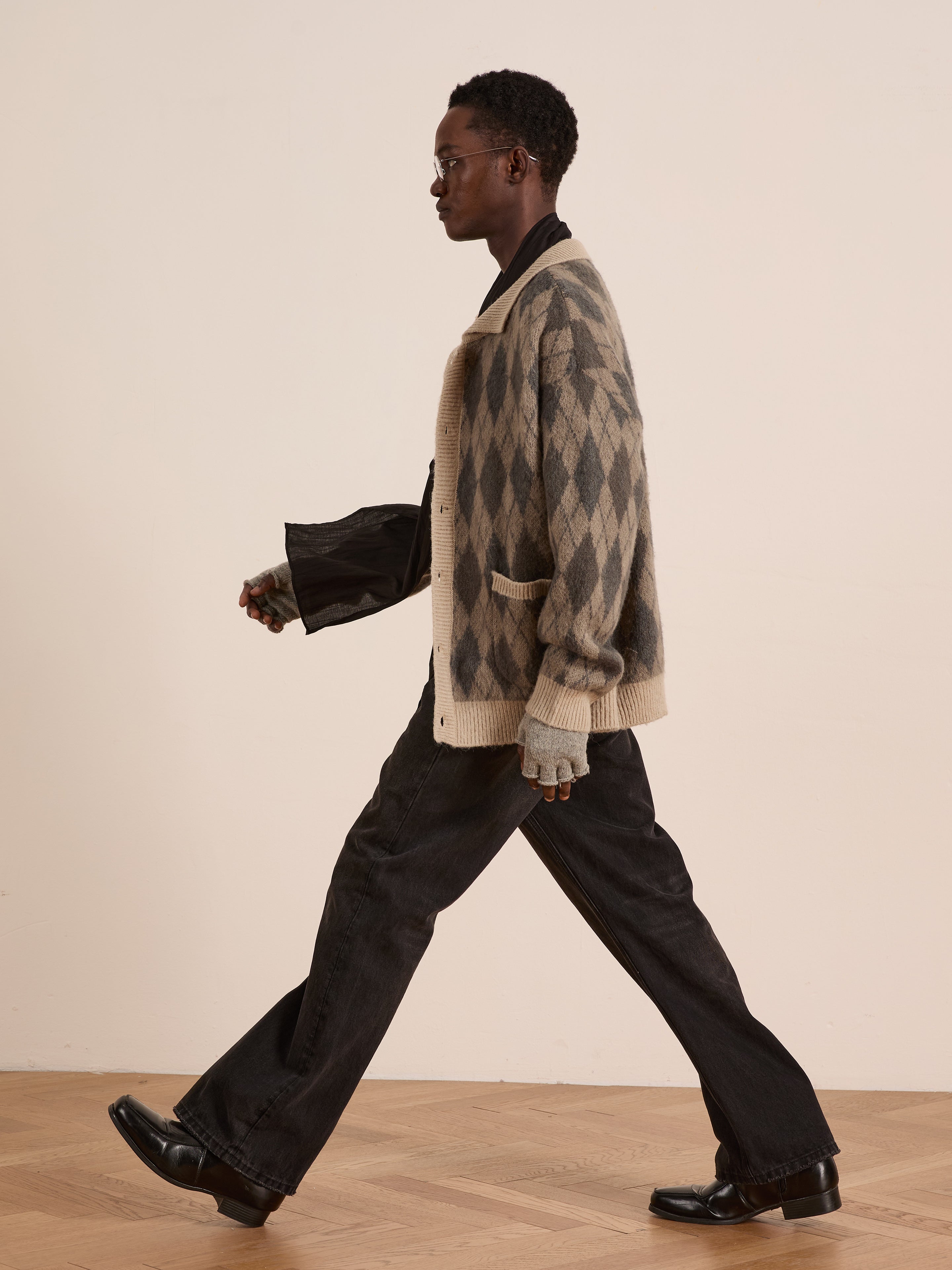 A person in glasses walks indoors against a plain light wall, wearing the FOUND Argyle Cardigan in beige and gray, paired with wide-leg black pants and black shoes.