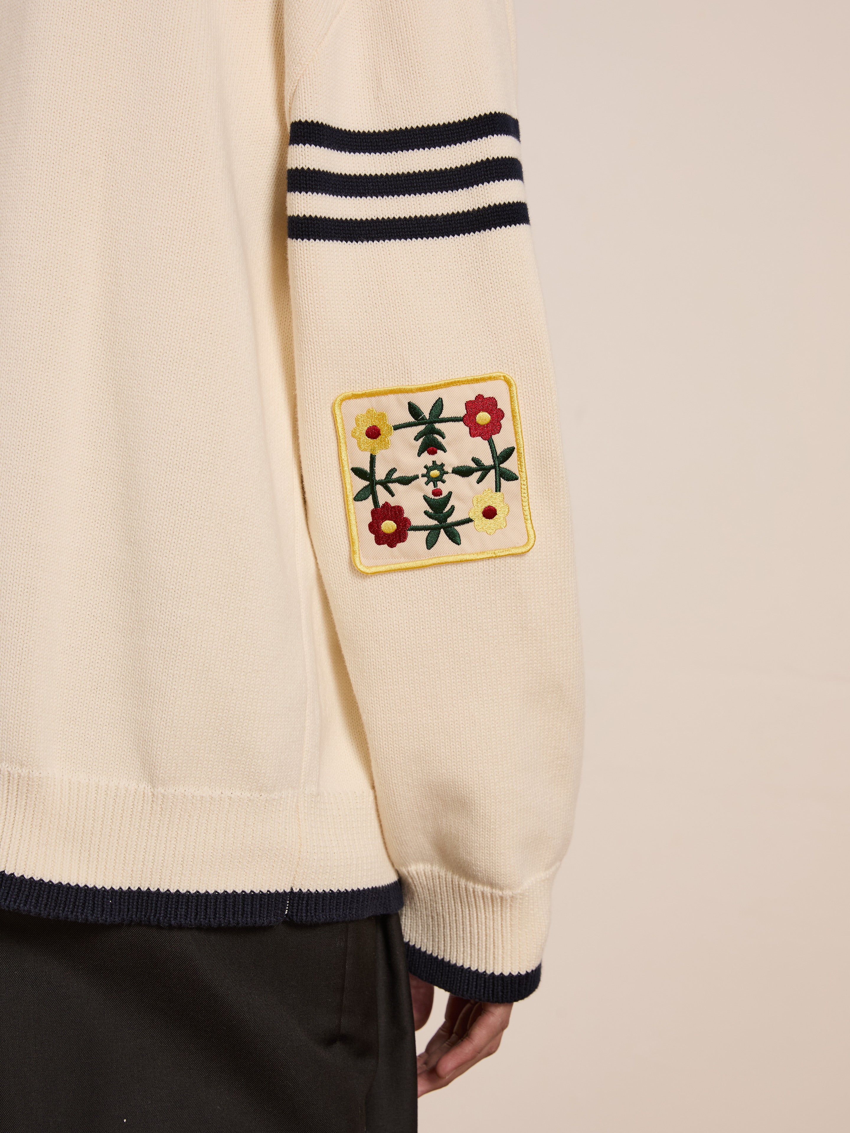 A close-up shows someone wearing the York Varsity Patch Collared Cardigan by FOUND—a cream sweater with black sleeve stripes and a floral patch, featuring red, yellow, and green embroidery on the elbow for a collegiate nostalgic look.