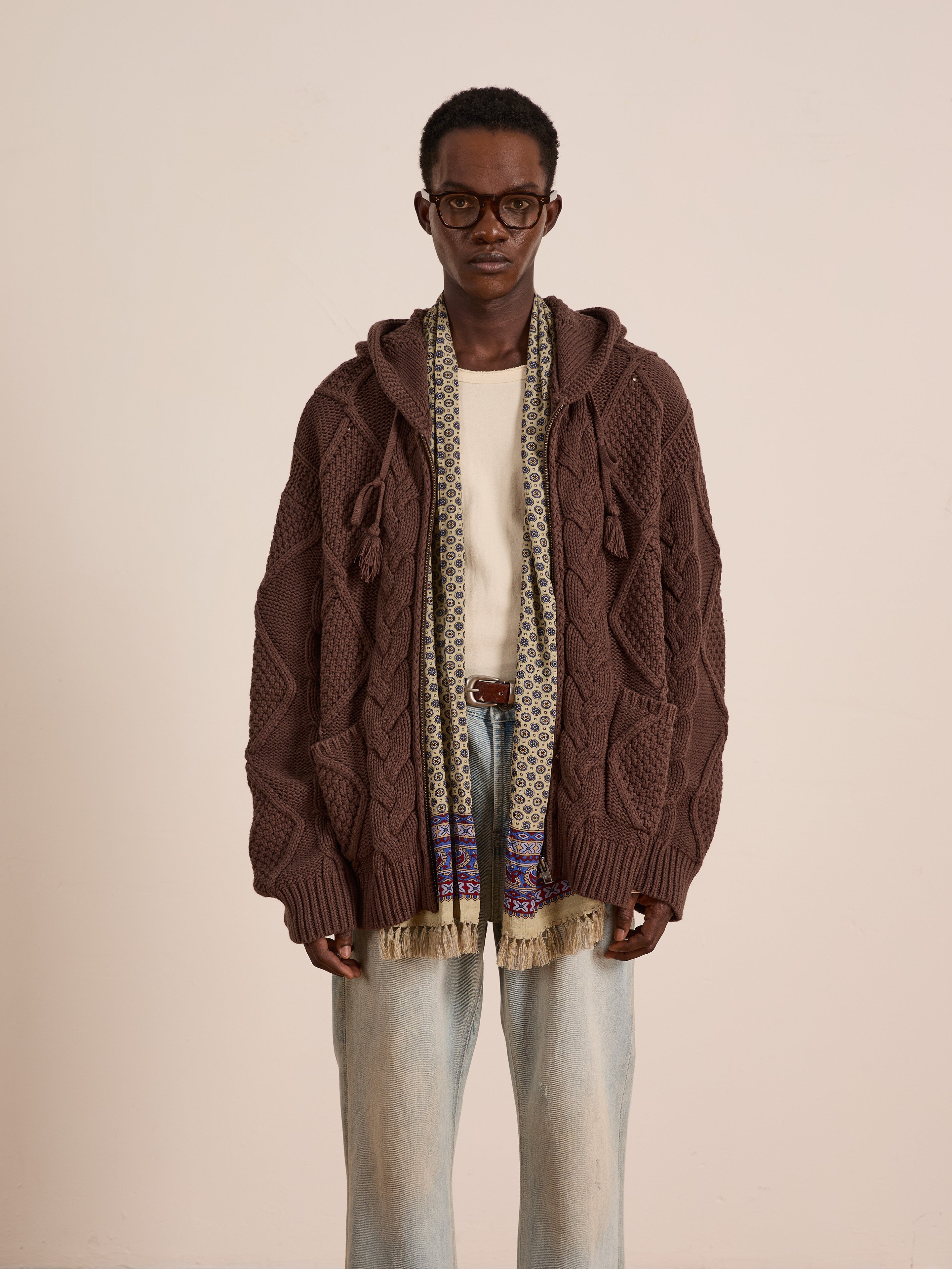 A person wears the FOUND Zip Up Cable Knit Hoodie in a relaxed unisex fit, styled with glasses, a cream shirt, light blue jeans, and a patterned scarf, set against a plain light background.