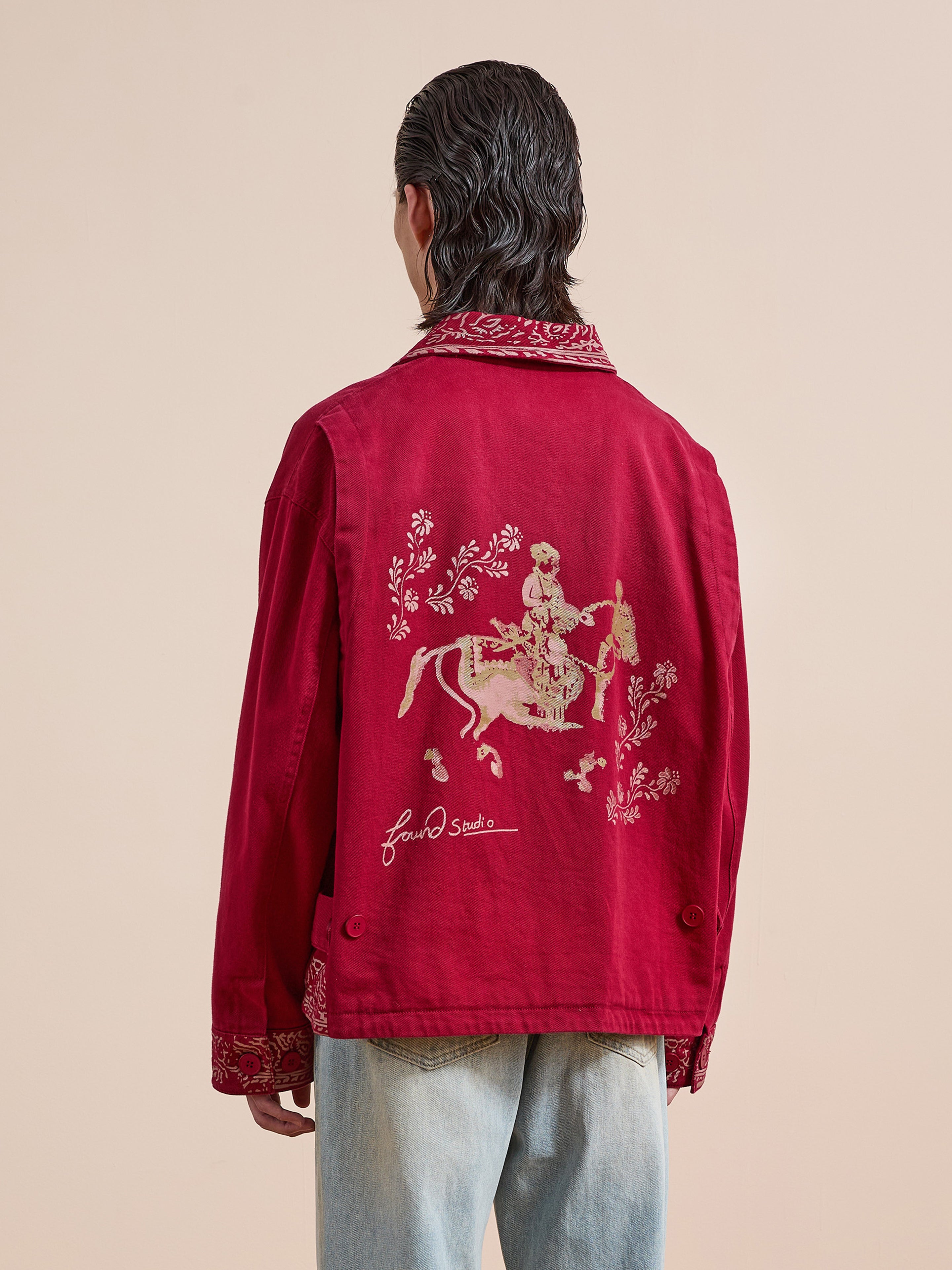 A person faces away, wearing Found’s Abhala Printed Work Jacket with a unisex relaxed fit, floral embroidery, and equestrian motifs on the back, paired with light blue jeans against a plain background.