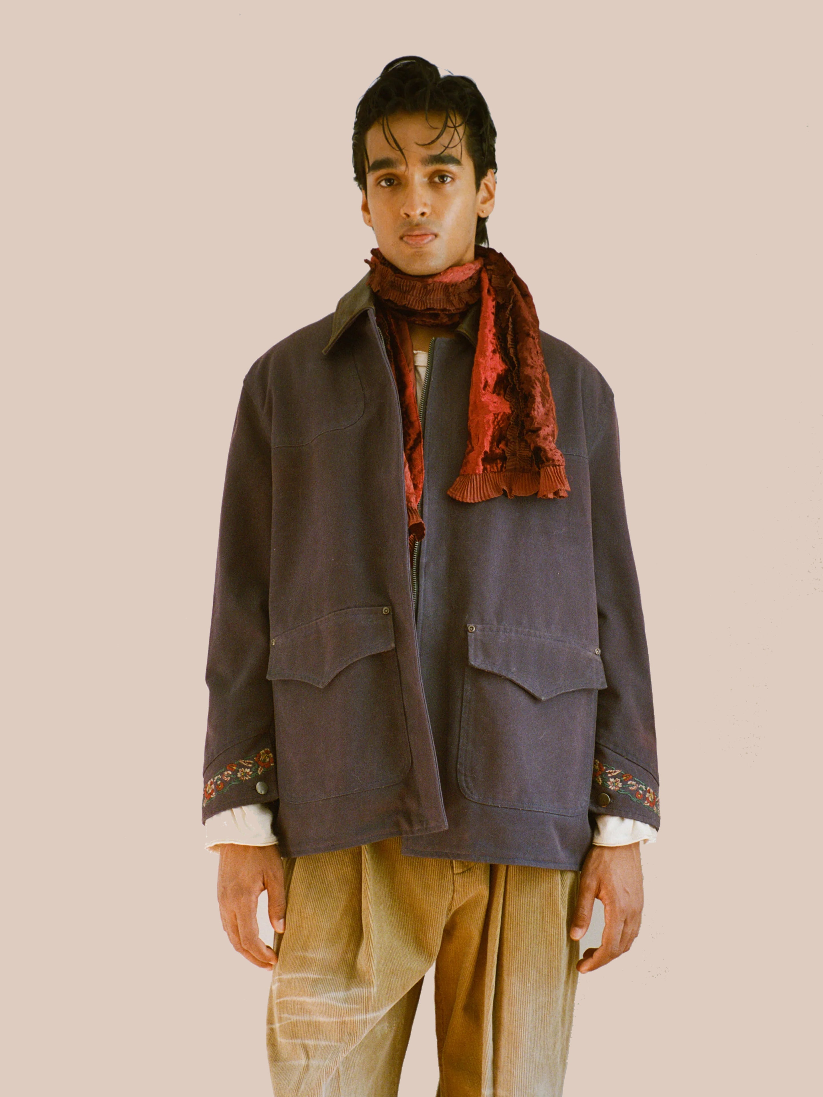 A person stands against a plain background wearing the Found Canvas Distressed Barn Jacket with a leather collar, embroidered cuffs, a red scarf, white shirt, and tan pants for a relaxed unisex fit.