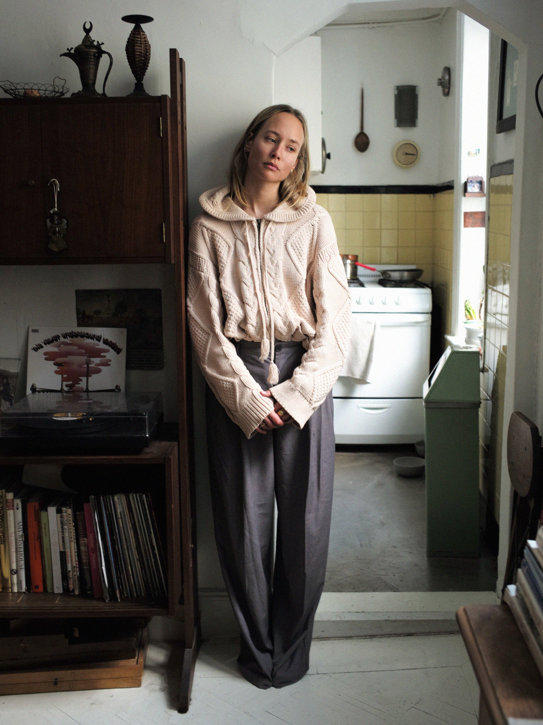 A person in a Zip Up Cable Knit Hoodie by Found, featuring a unisex relaxed fit, stands against a wall in a cozy kitchen with a record player and books.