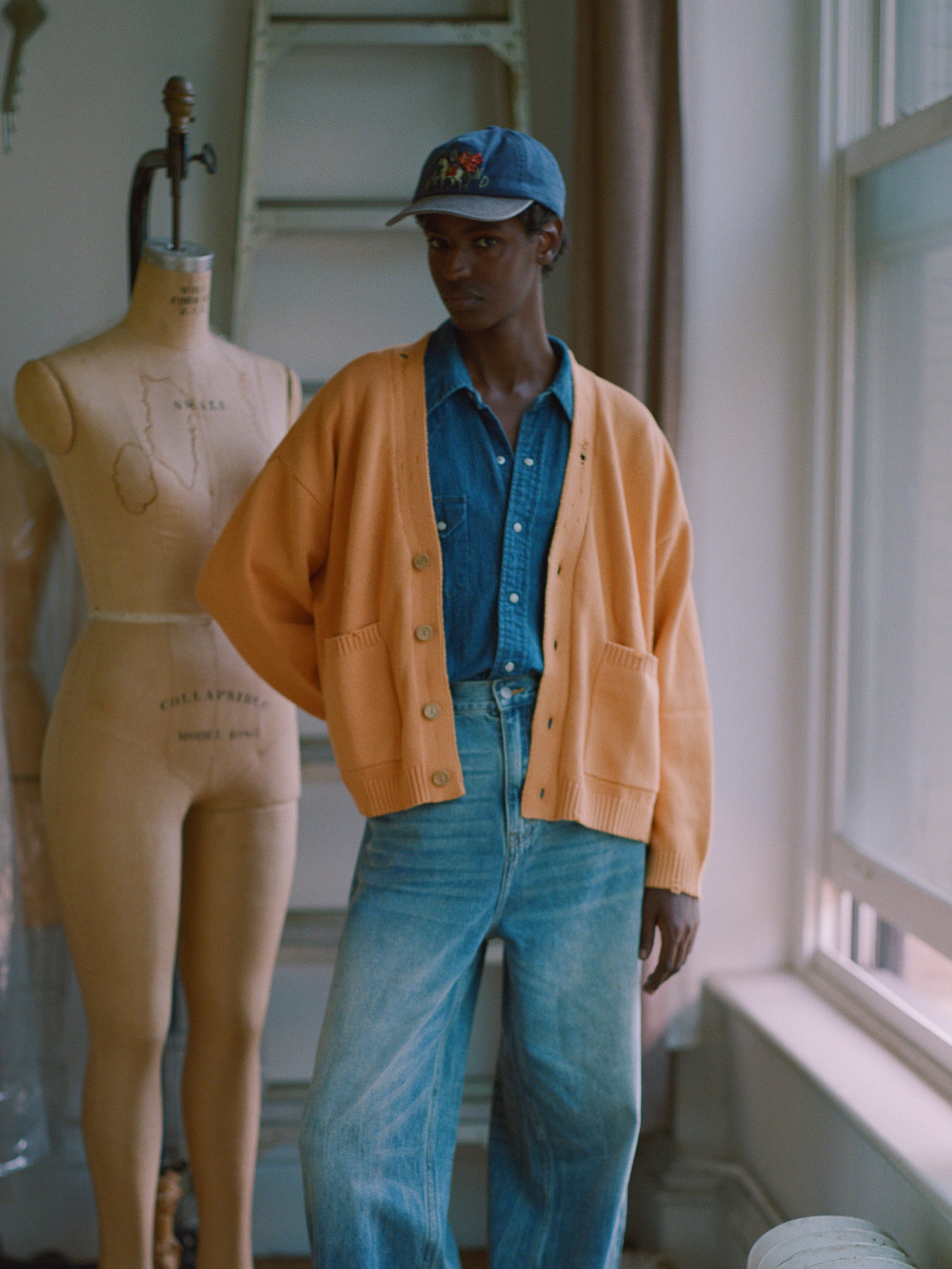 A man in a Cadmium Distressed Cardigan by Found with chunky ribbing standing next to a mannequin.