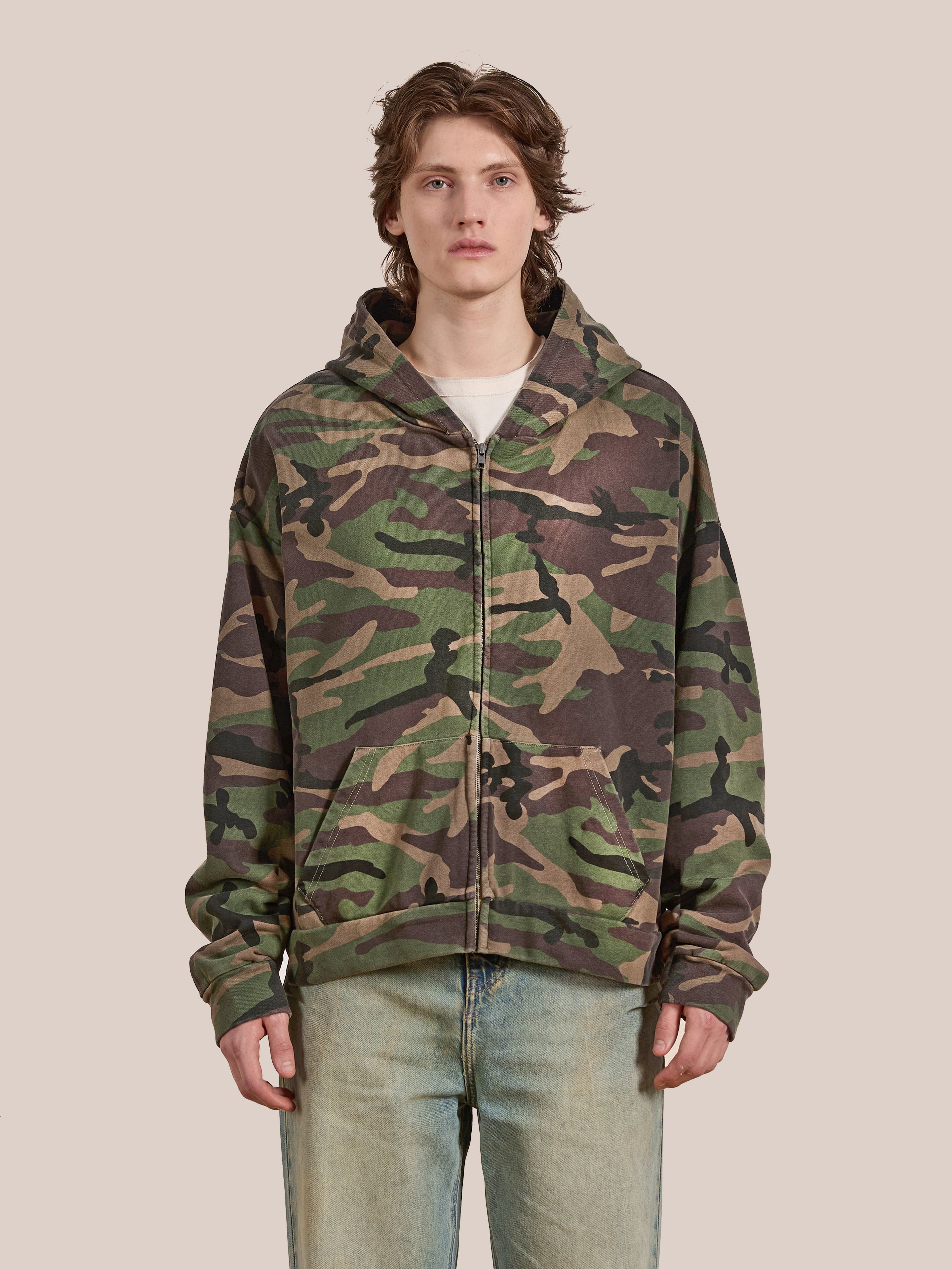 A person stands against a plain background wearing the Washed Camouflage Zip Hoodie by FOUND, paired with light blue jeans.