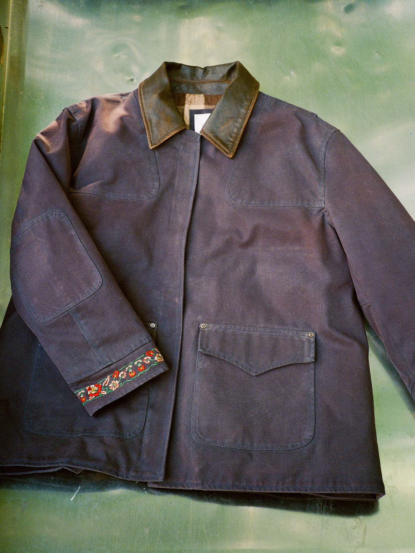 The Found Canvas Distressed Barn Jacket is a navy, unisex jacket with a relaxed fit, large front pockets, a distressed leather collar, and floral embroidery on one cuff, shown laid flat on a green surface.