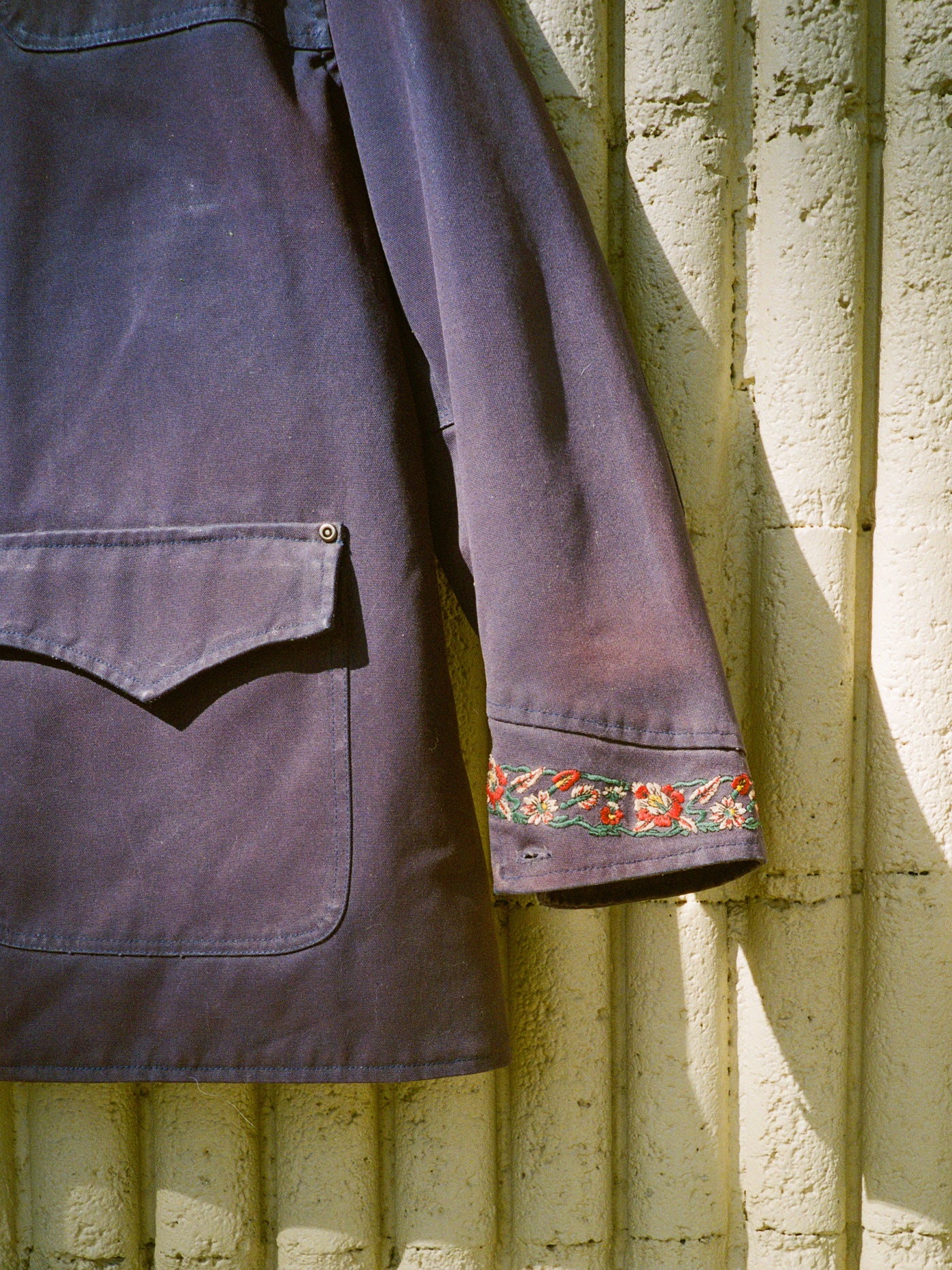 The Found Canvas Distressed Barn Jacket, a purple unisex relaxed fit with a large front pocket, distressed leather collar, and floral sleeve embroidery, hangs against a textured light wall in sunlight.