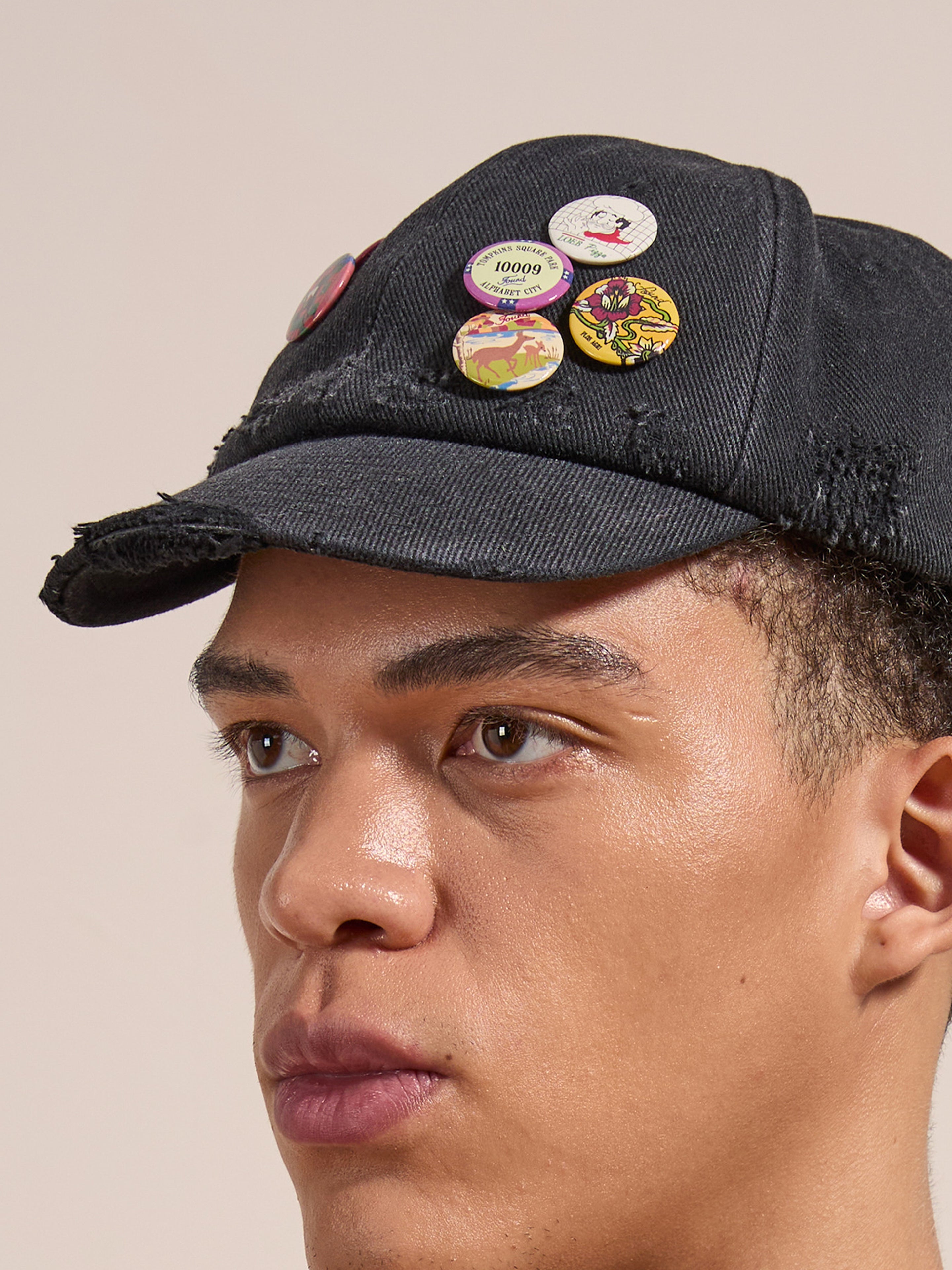 A person wears the FOUND Distressed Pin Cap, a black cotton 6-panel hat with a worn-in look, adorned with multiple colorful vintage pins in different designs.