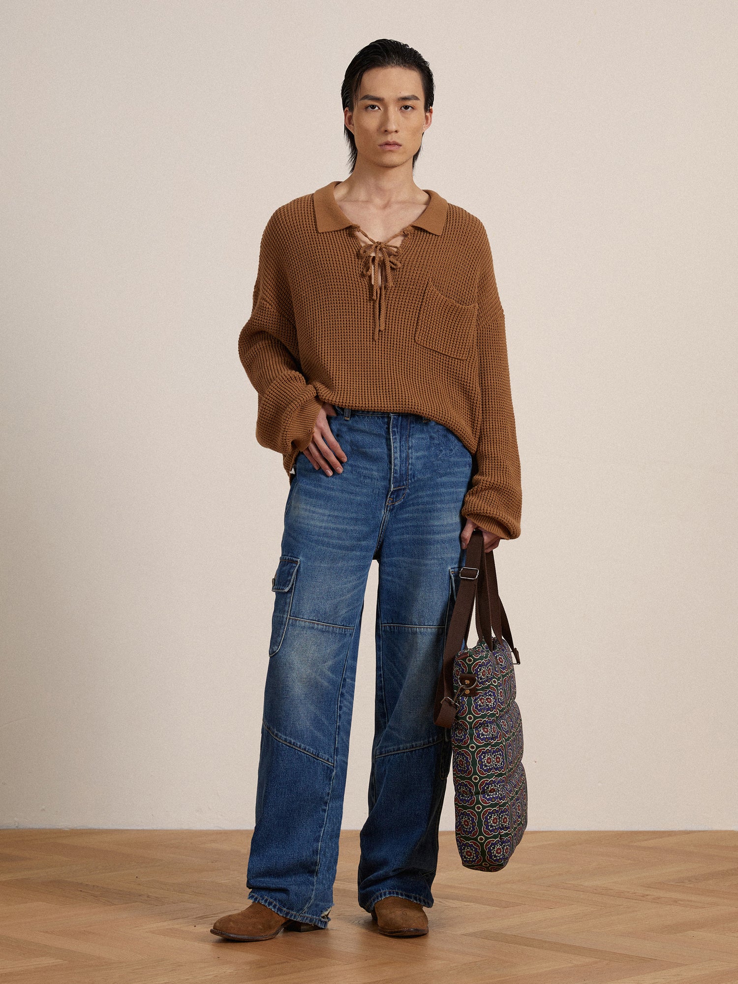 A person in a brown knitted top and Found Siwa Cargo Paneled Jeans, holding a patterned bag, stands in a neutral-toned room.