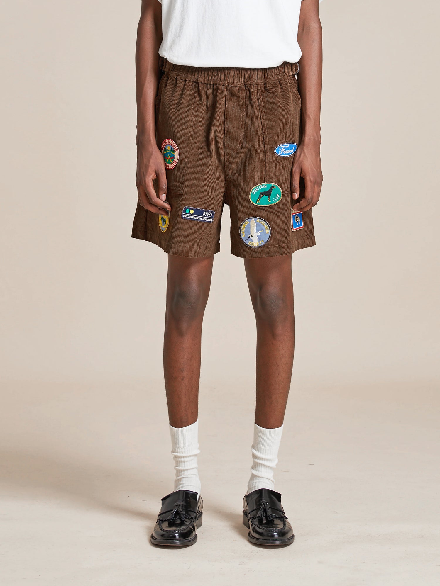 A person wearing Found Canoe Multi Patch Corduroy Shorts with colorful embroidered patches, white socks, and black shoes, standing against a neutral background. Only the lower half of the body is visible.