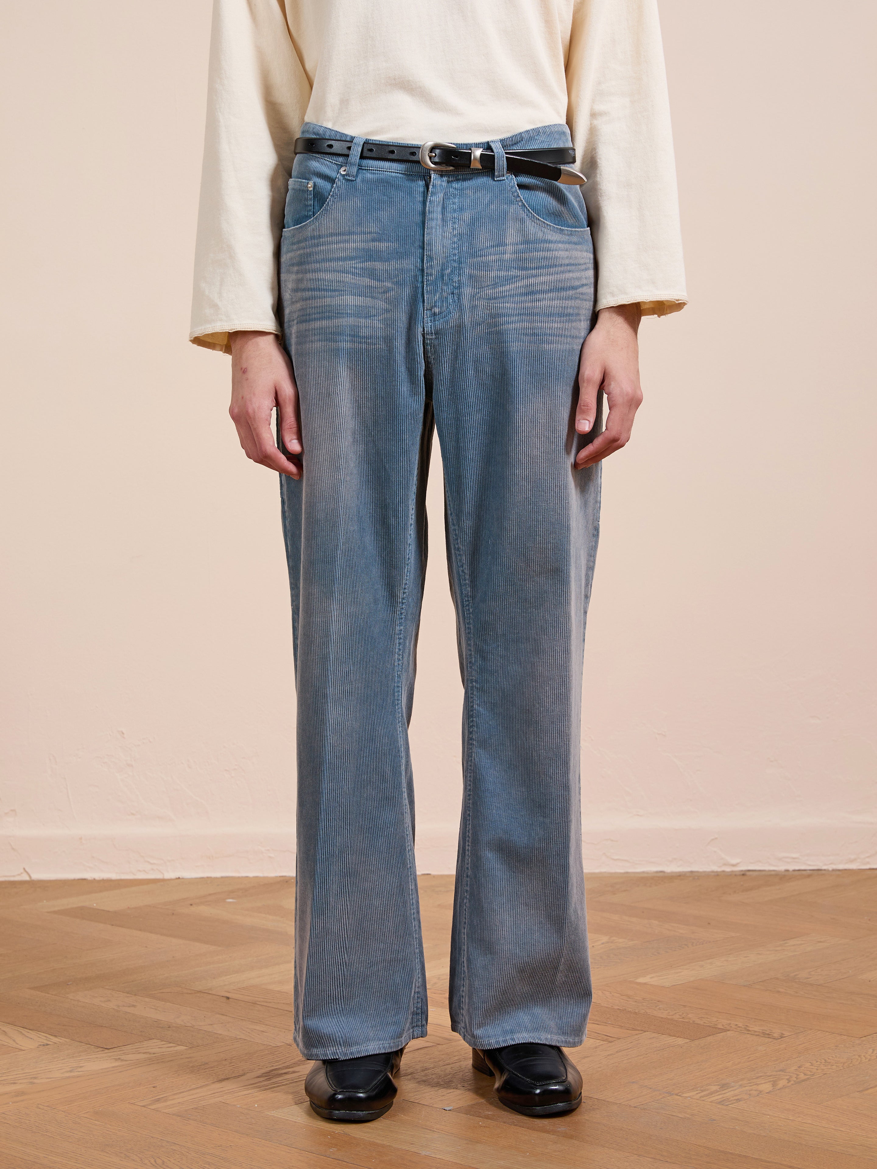 A person wears FOUND Corduroy Flare Work Pants in vintage blue, styled with a cream long-sleeve shirt, black belt, and black shoes, standing on a wooden floor against a beige wall.