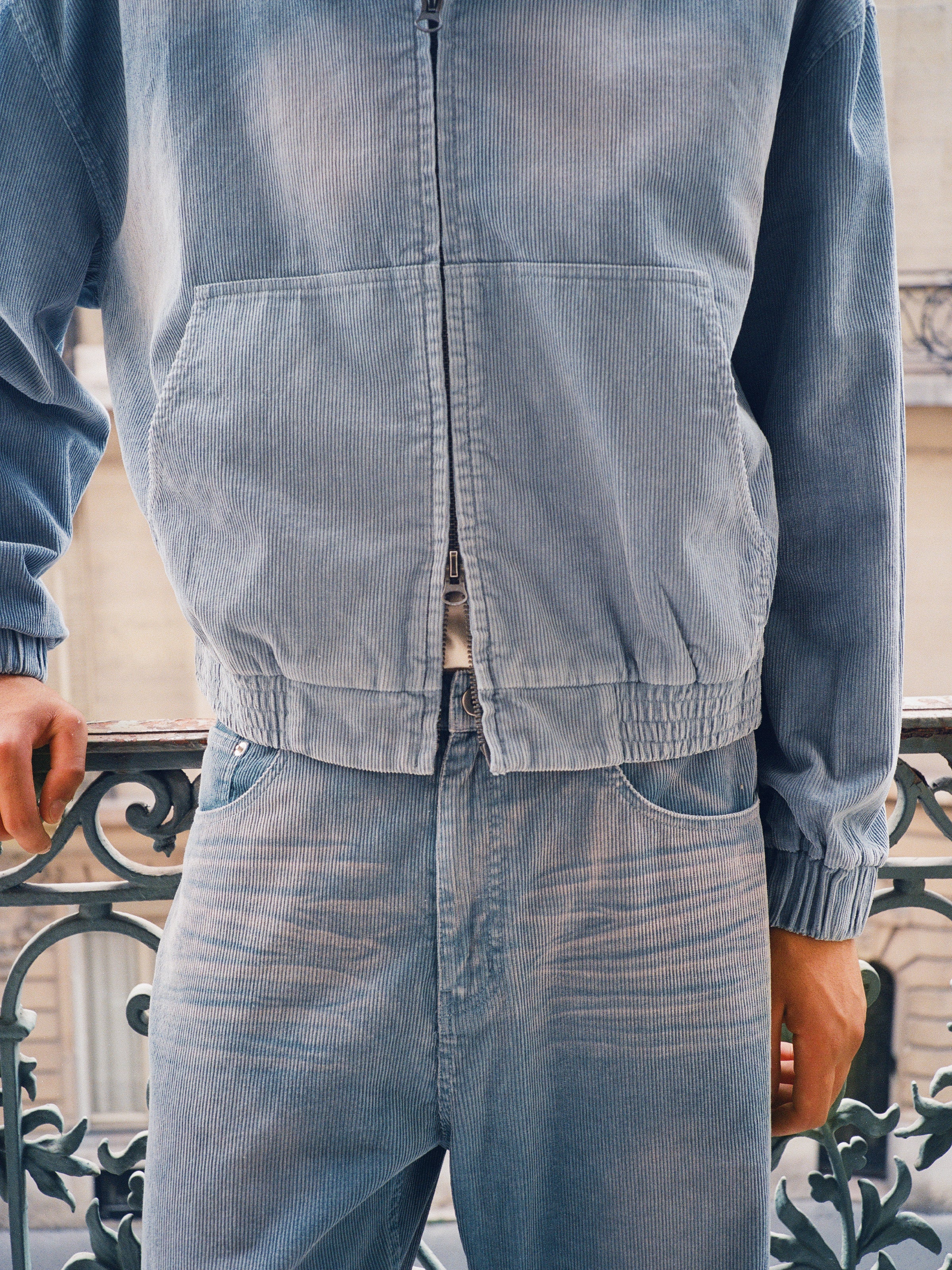 A person wearing FOUND Corduroy Flare Work Pants and a vintage blue corduroy jacket stands by a decorative metal railing, highlighting the pants' relaxed fit.