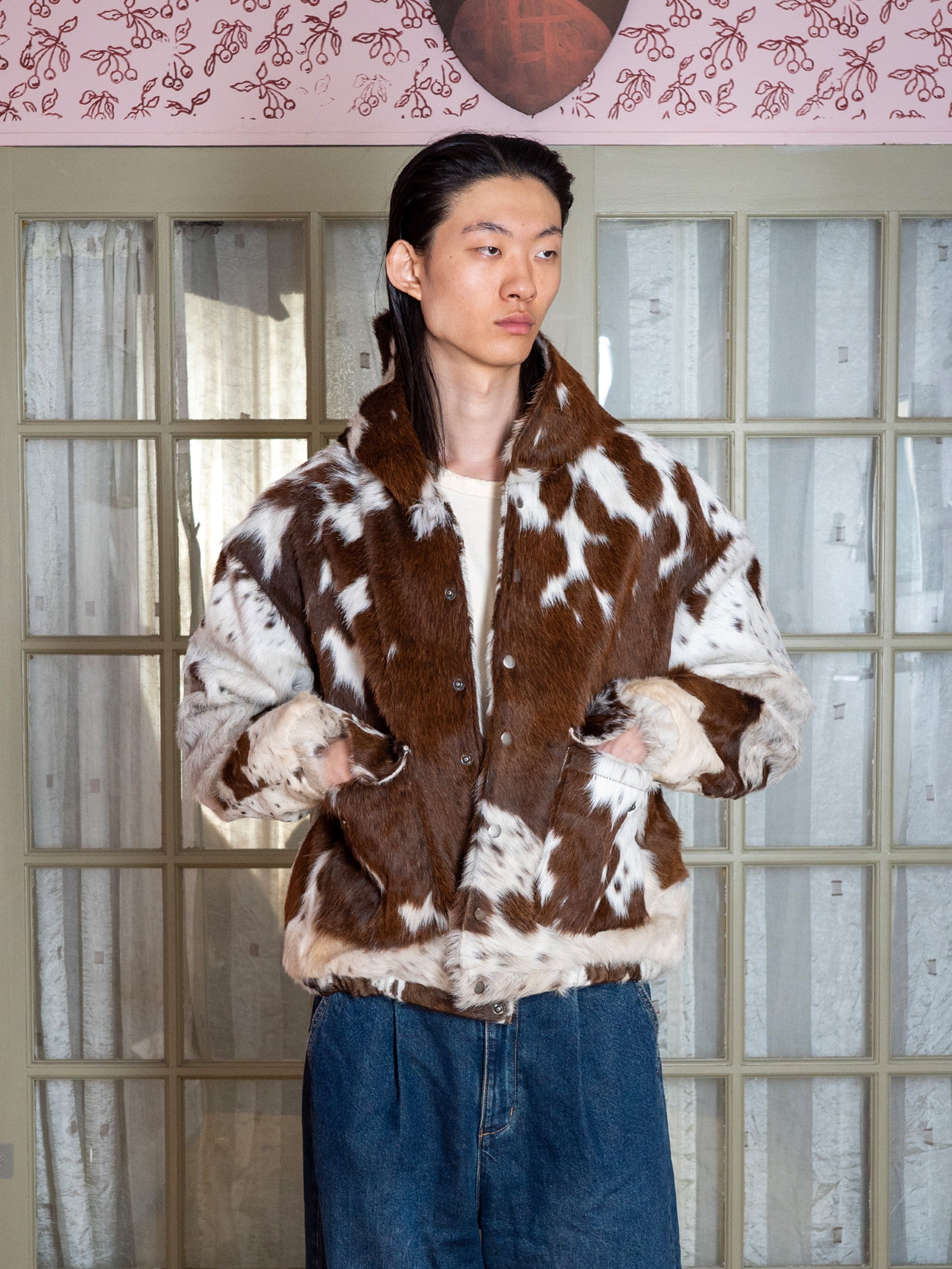 A person wears the FOUND Cowhide Drover Hoodie, featuring a brown and white cow print, paired with unisex relaxed-fit blue jeans, standing indoors by glass-paneled doors.
