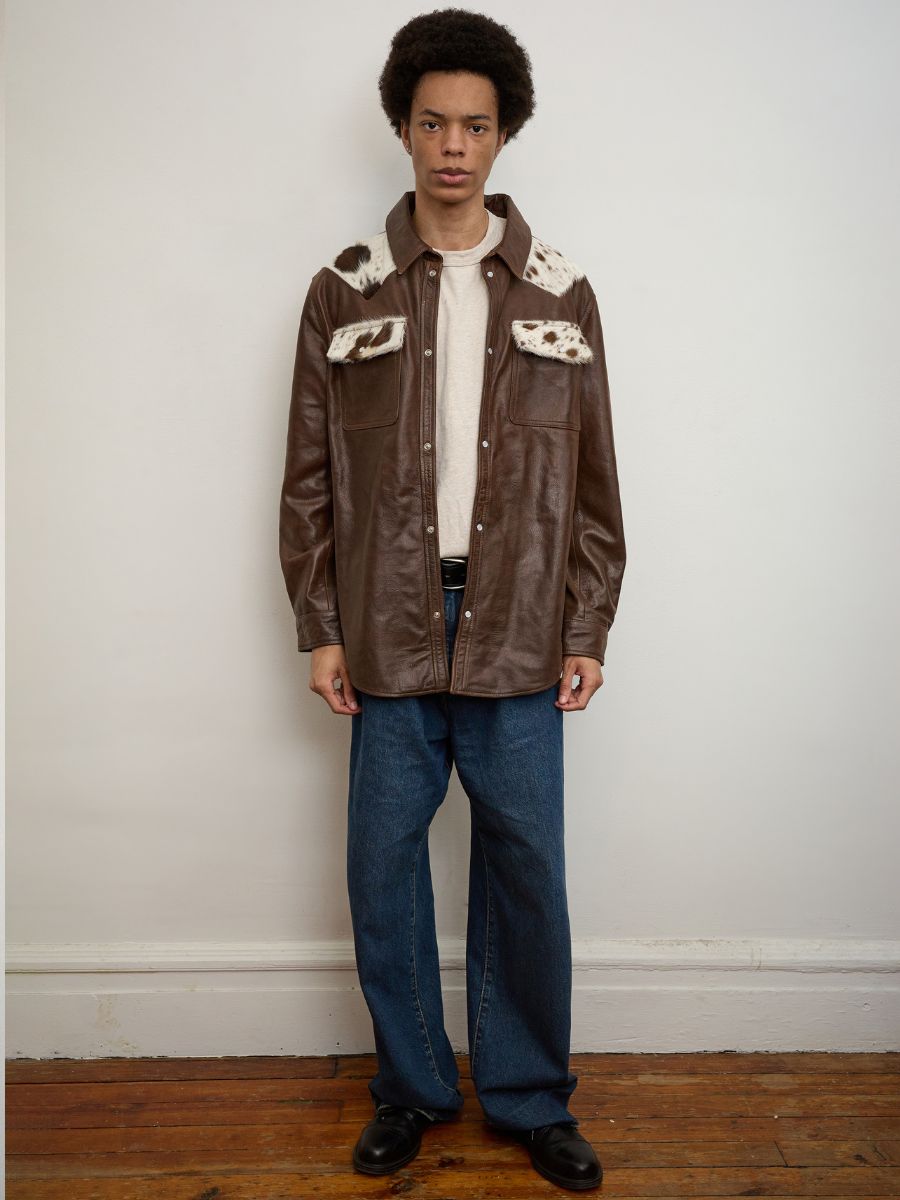 A person stands indoors against a white wall and wooden floor, wearing blue jeans, black shoes, and the FOUND Distressed Leather Cowhide Contrast Western Shirt.