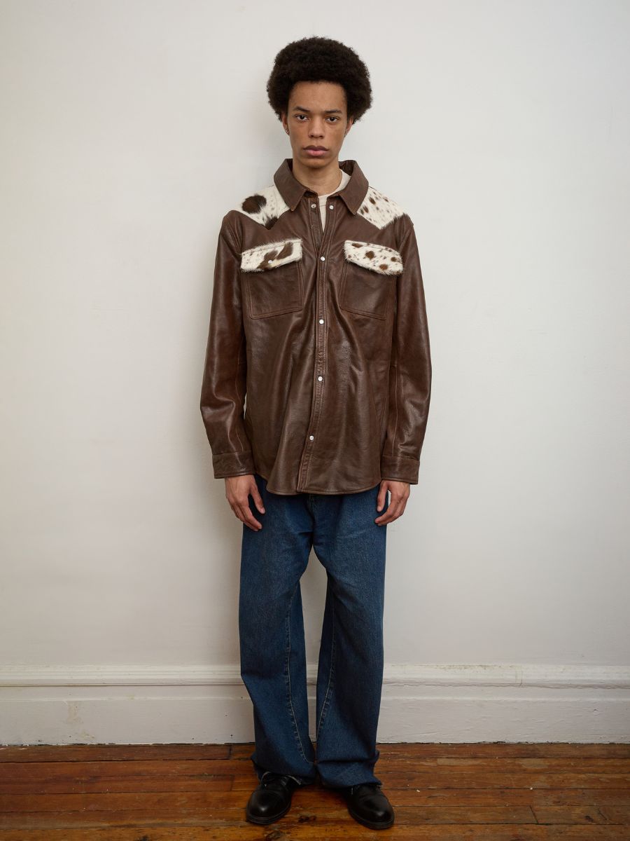 A person stands indoors against a white wall, wearing the FOUND Distressed Leather Cowhide Contrast Western Shirt, paired with blue jeans and black shoes.