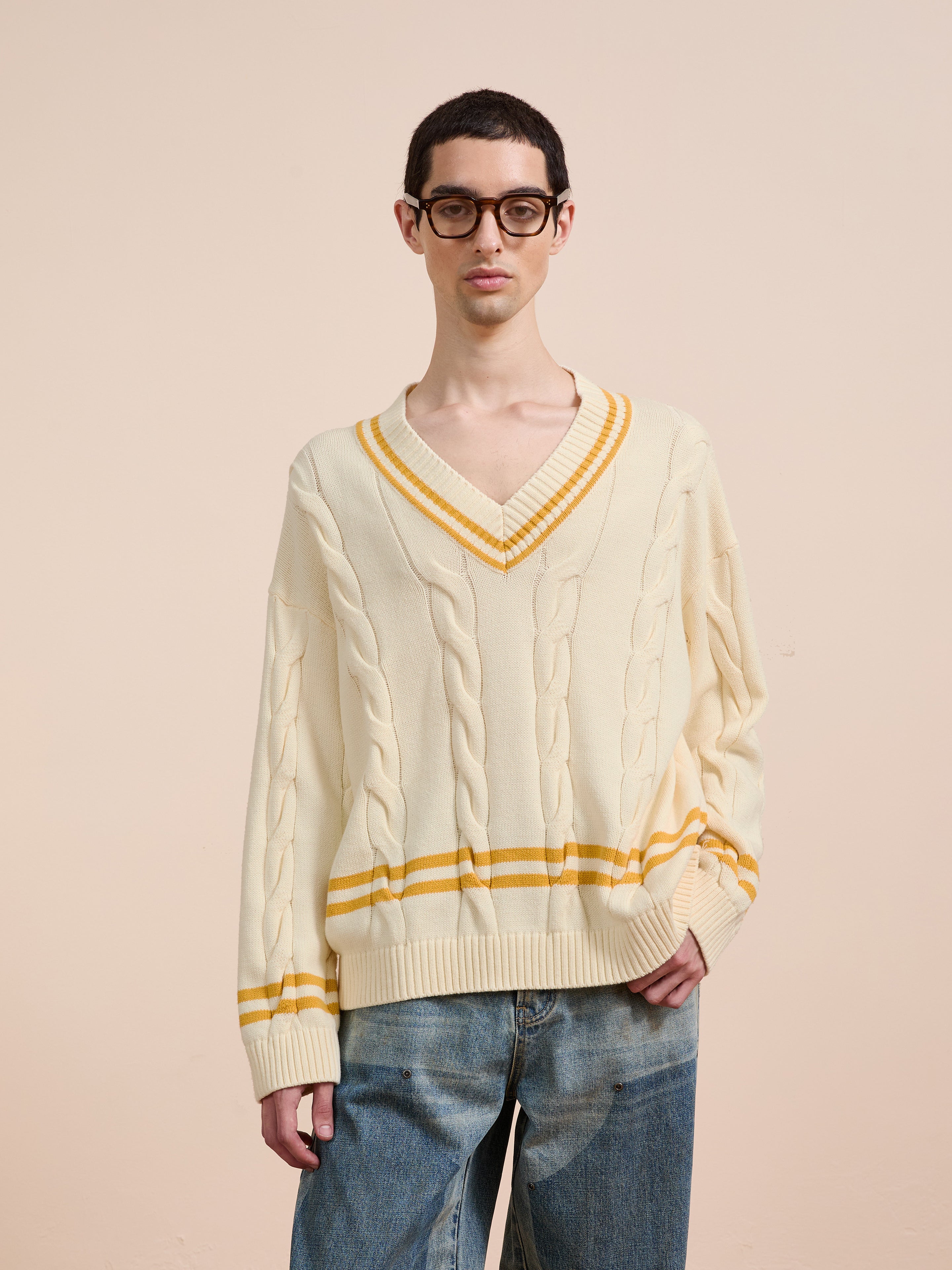 A person in blue jeans and glasses wears the FOUND V-Neck Cricket Sweater with yellow stripes, standing against a plain beige background.