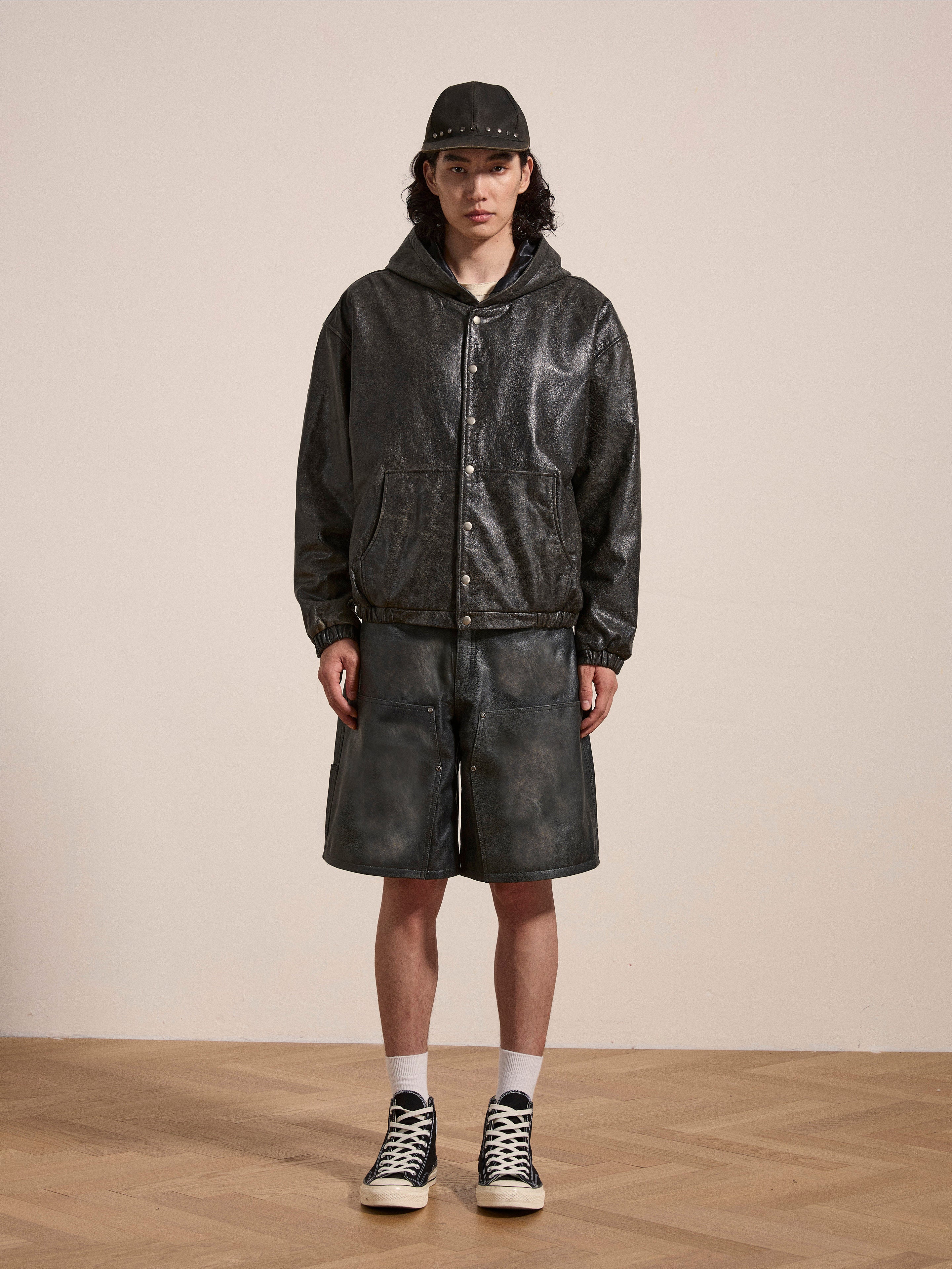 A person wearing a dark Distressed Genuine Leather Hoodie by Found, matching leather shorts, a black cap, black high-top sneakers, and white socks stands in front of a neutral background.