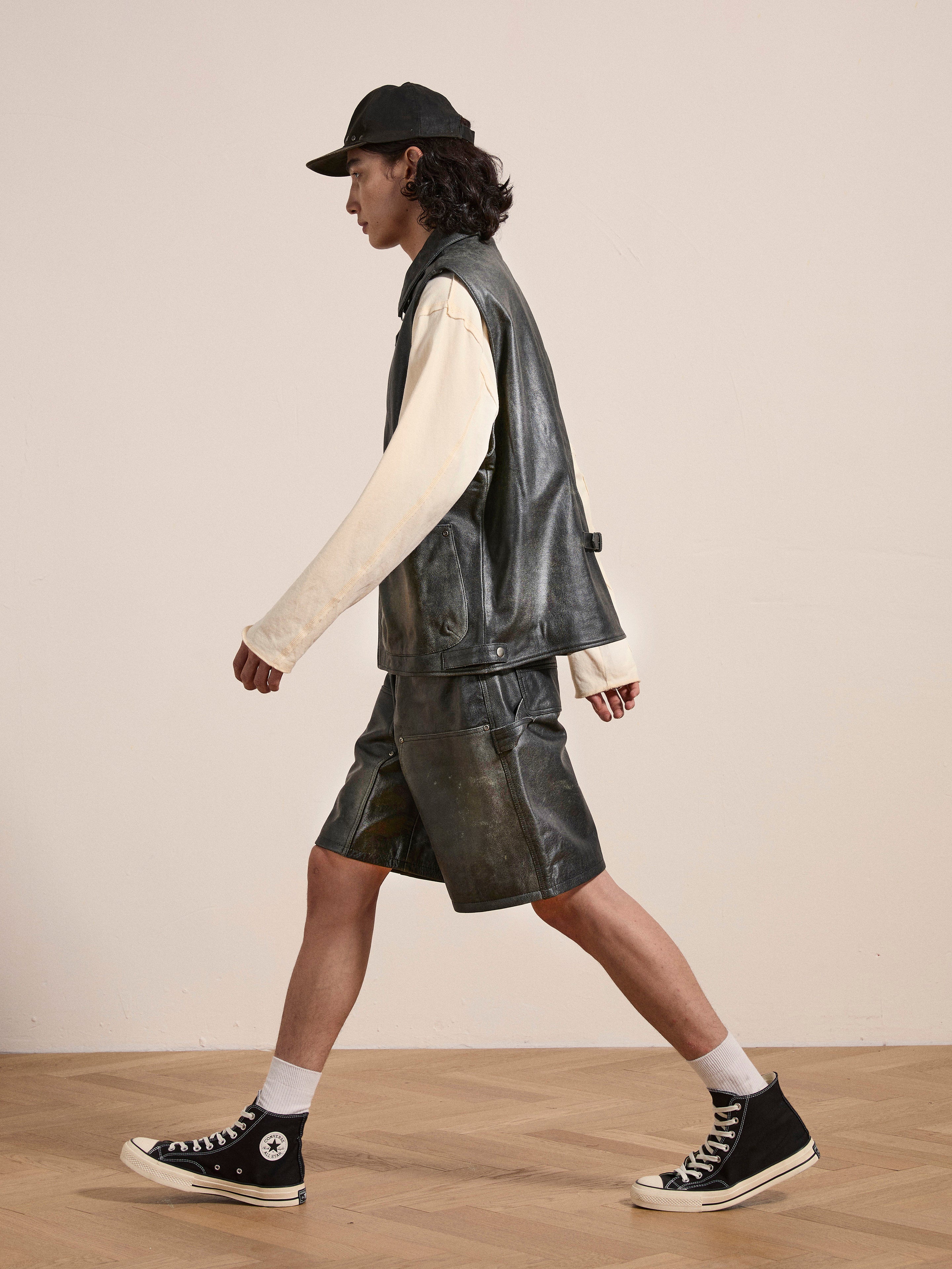 A person wearing a black cap, sleeveless leather jacket, white long-sleeve shirt, Found Distressed Genuine Leather Carpenter Shorts, white socks, and black high-top sneakers walks on a wooden floor.