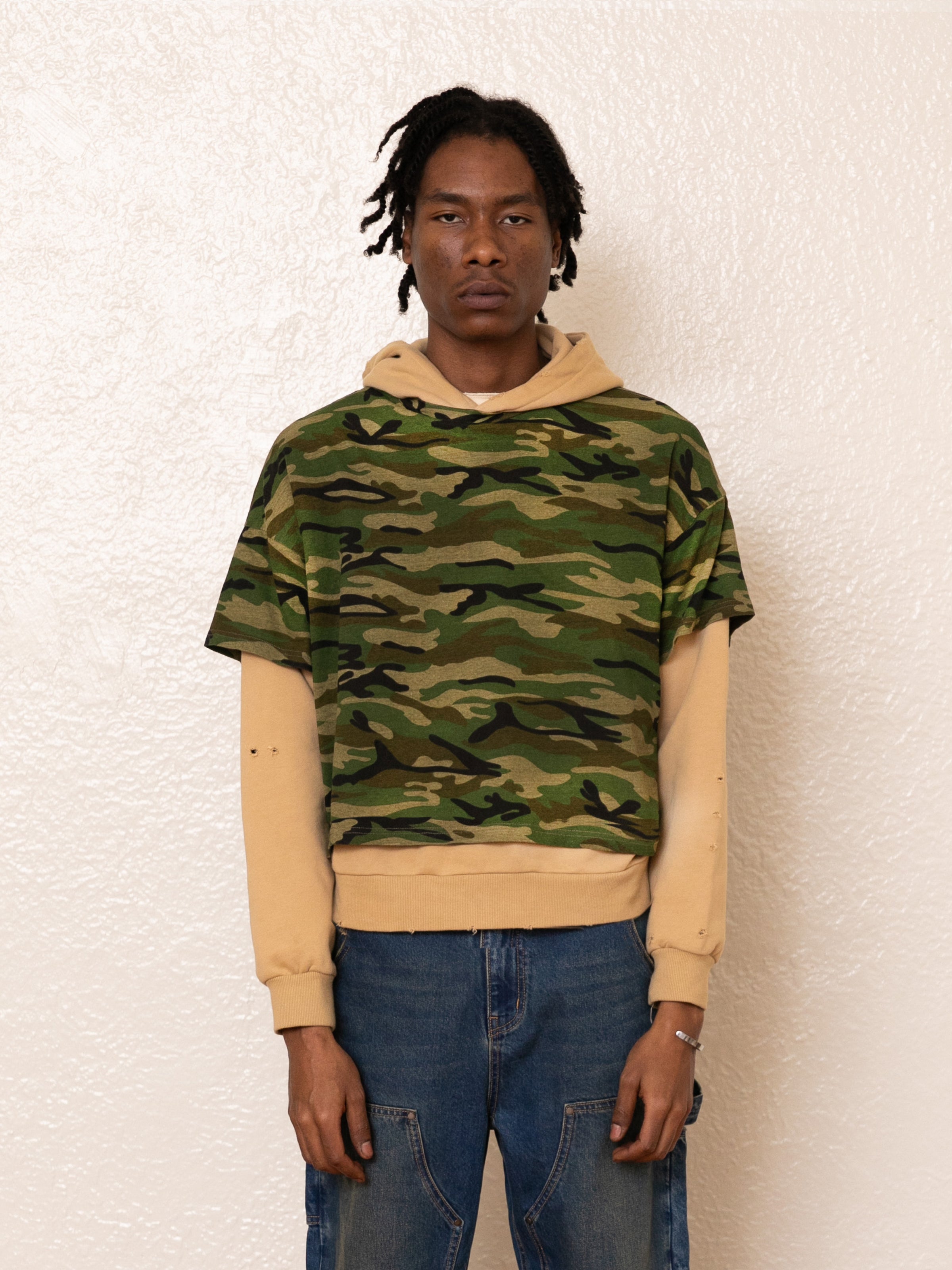 A person with dreadlocks sports a vintage style, donning FOUND's Double Layer Hoodie in french terry cotton over a camo t-shirt and blue jeans, standing against a textured white wall.