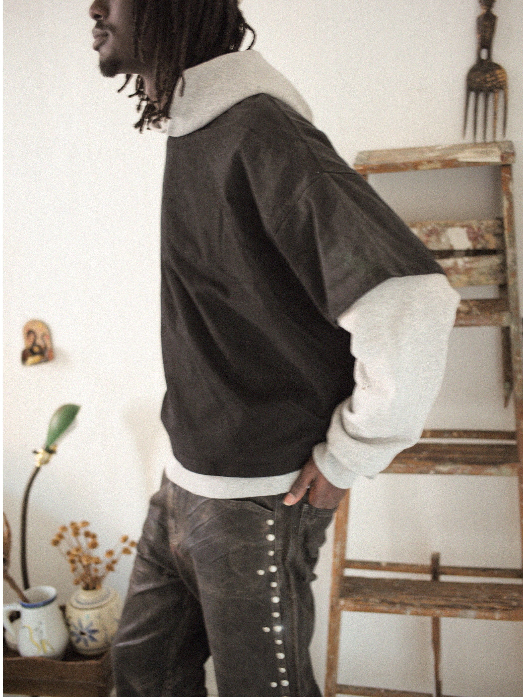 A person in a black and gray outfit stands sideways indoors beside a wooden ladder and decorative items, wearing the stylish Double Layer Hoodie by Found, which adds dimension to their look.