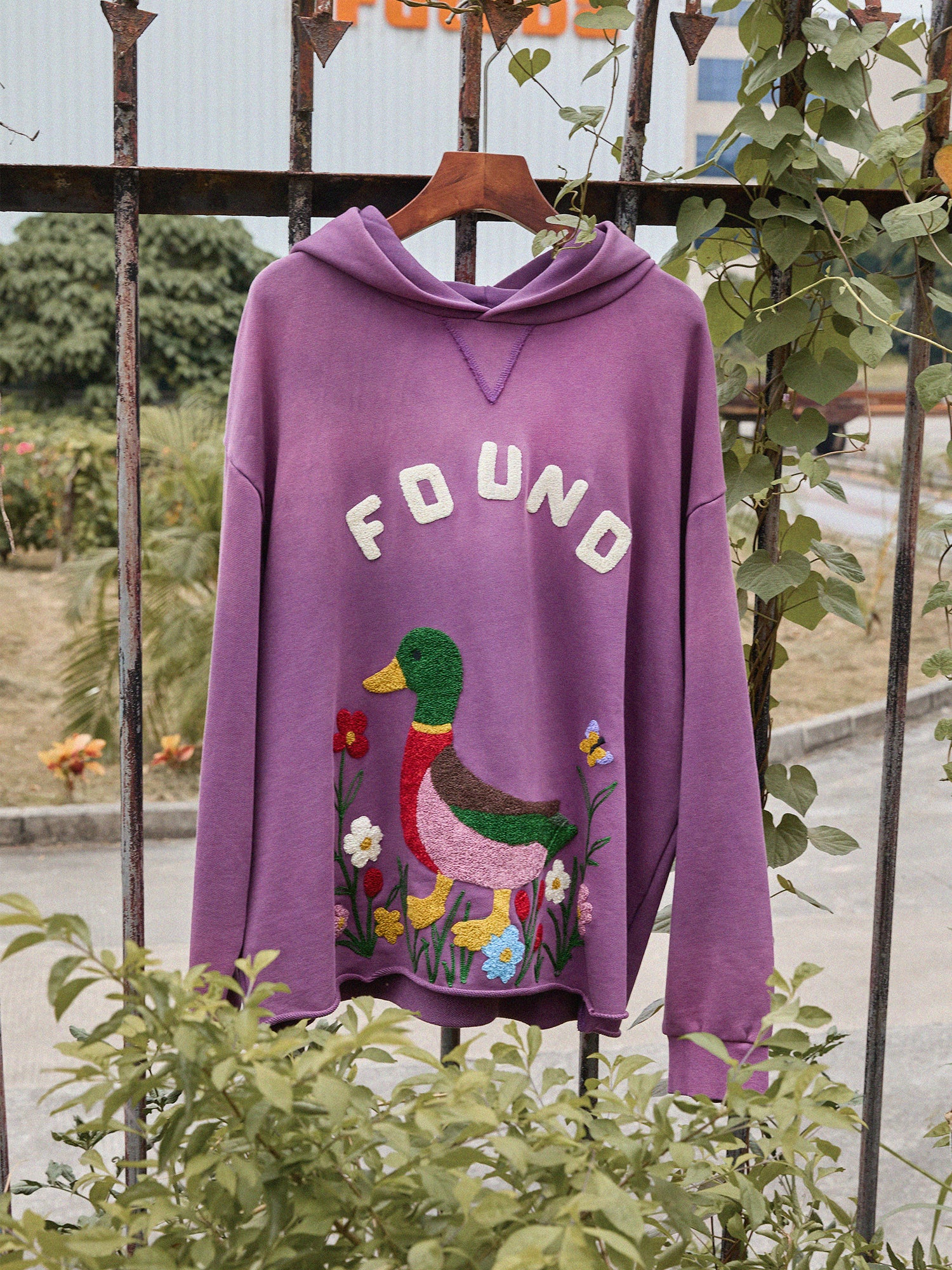 The FOUND Embroidered Wildlife Hoodie in purple, with an embroidered duck and flowers and "FOUND" above, hangs on a metal fence with lush greenery behind.