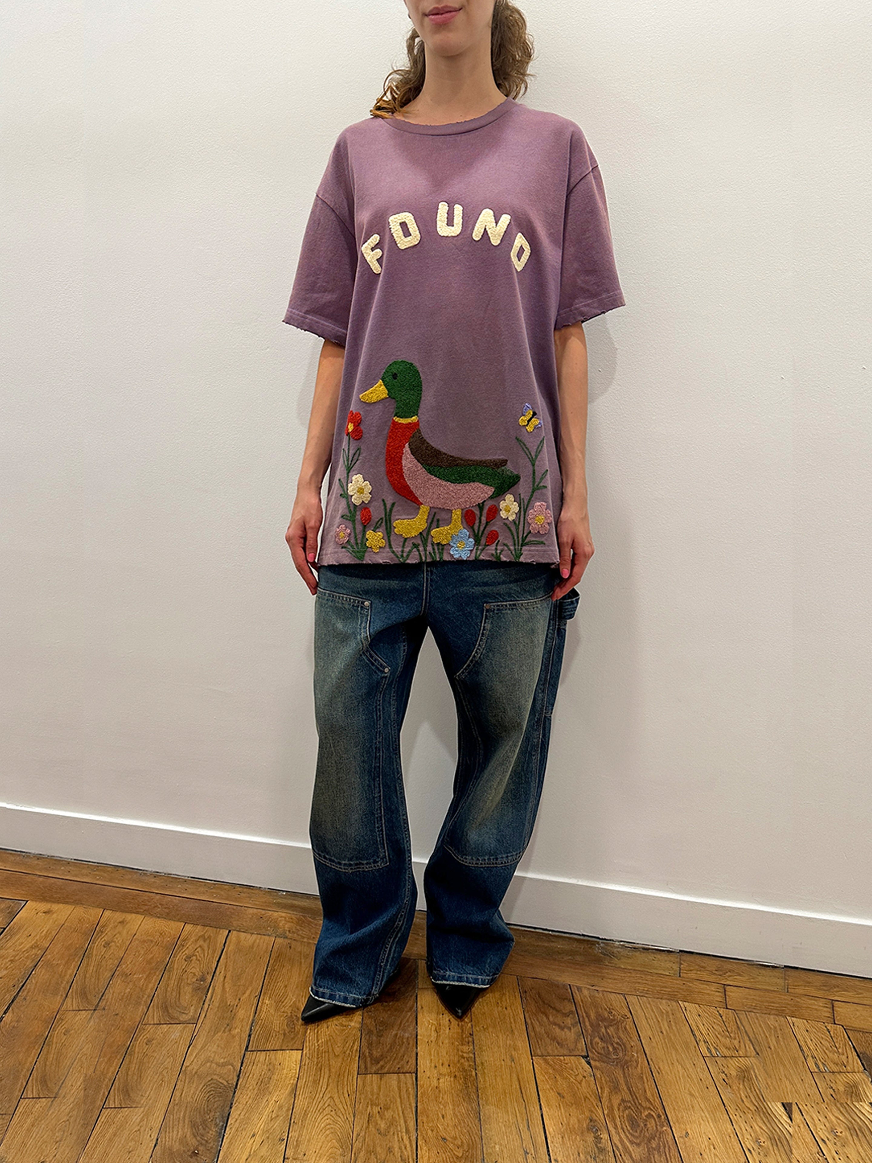 Indoors on a wooden floor, a person wears FOUND's Embroidered Wildlife Tee—purple with chenille duck and flower details—paired with loose blue jeans, standing against a white wall.