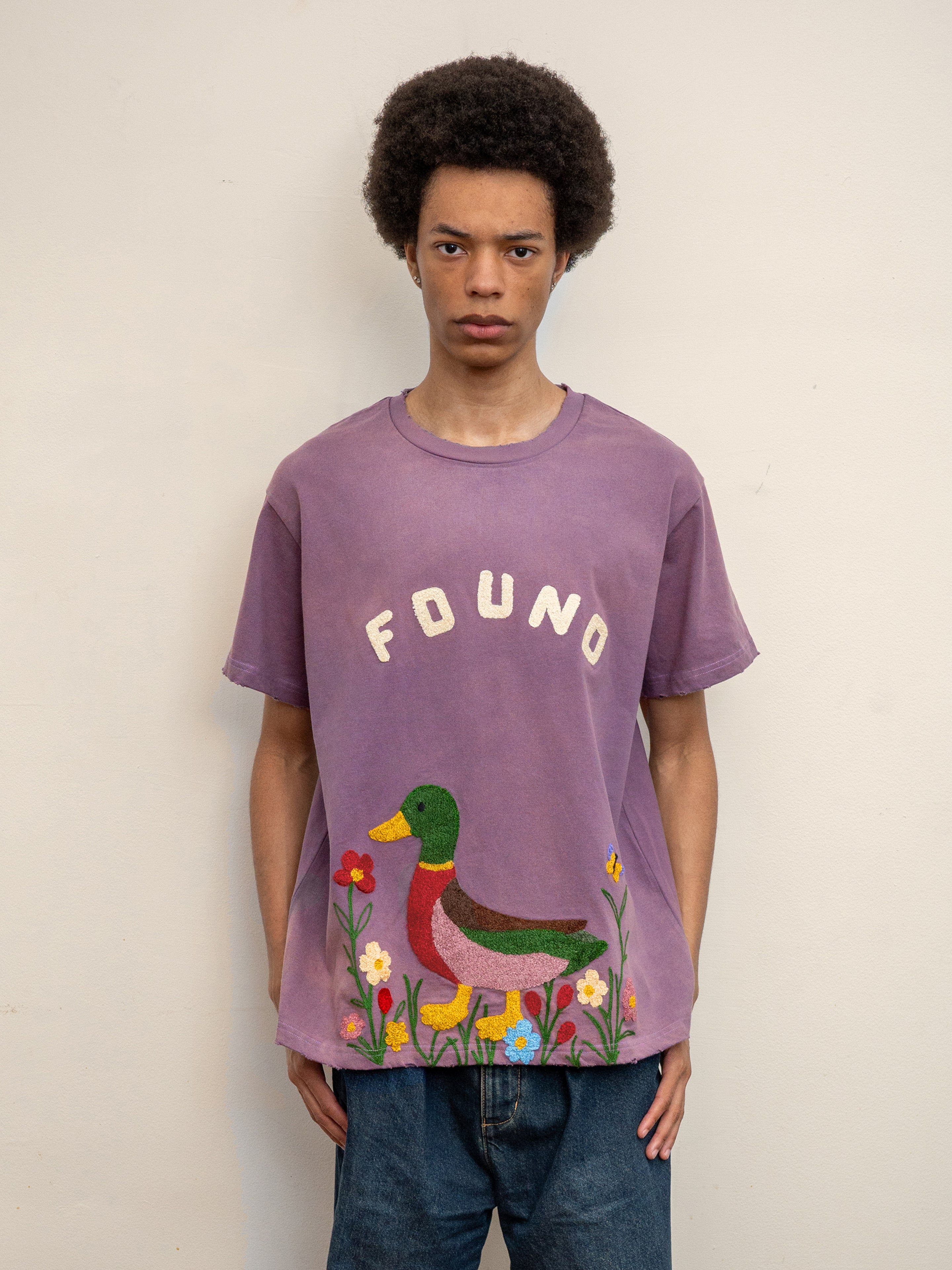 A person stands against a plain wall wearing the FOUND Embroidered Wildlife Tee, featuring "FOUND" text and a colorful duck and flower chenille embroidery on the front.