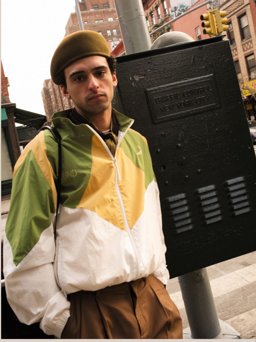 A person wearing a beret and the FOUND Legacy Cricket Track Jacket in green and yellow stands on a city street near a traffic control box labeled "New York City.