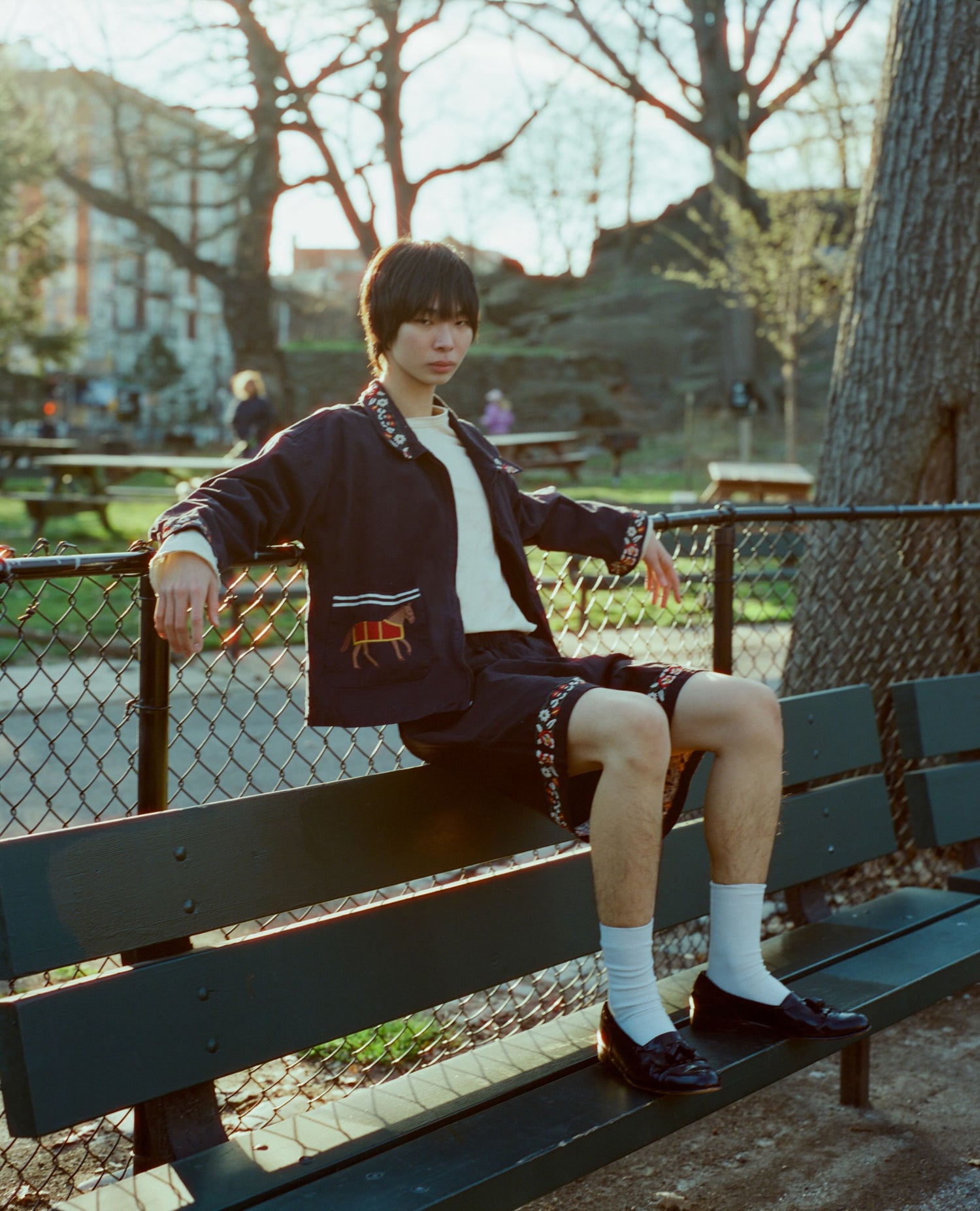 A young man sitting on a park bench, wearing a Found Horse Equine Work Jacket and shorts, with a relaxed posture and a slight smile.