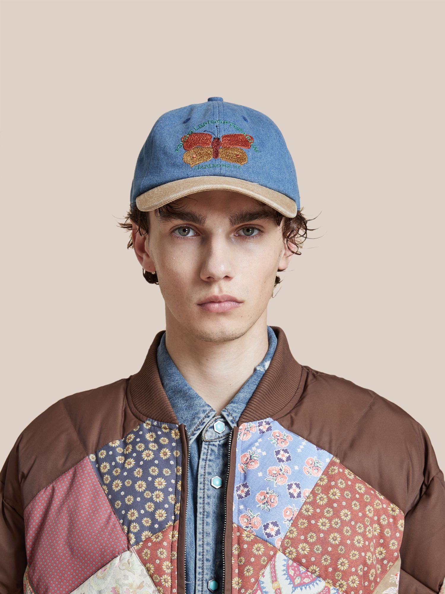Young man wearing a patchwork jacket and a Butterfly House Denim Cap with a corduroy brim, embroidered with a butterfly, posing against a light pink background.