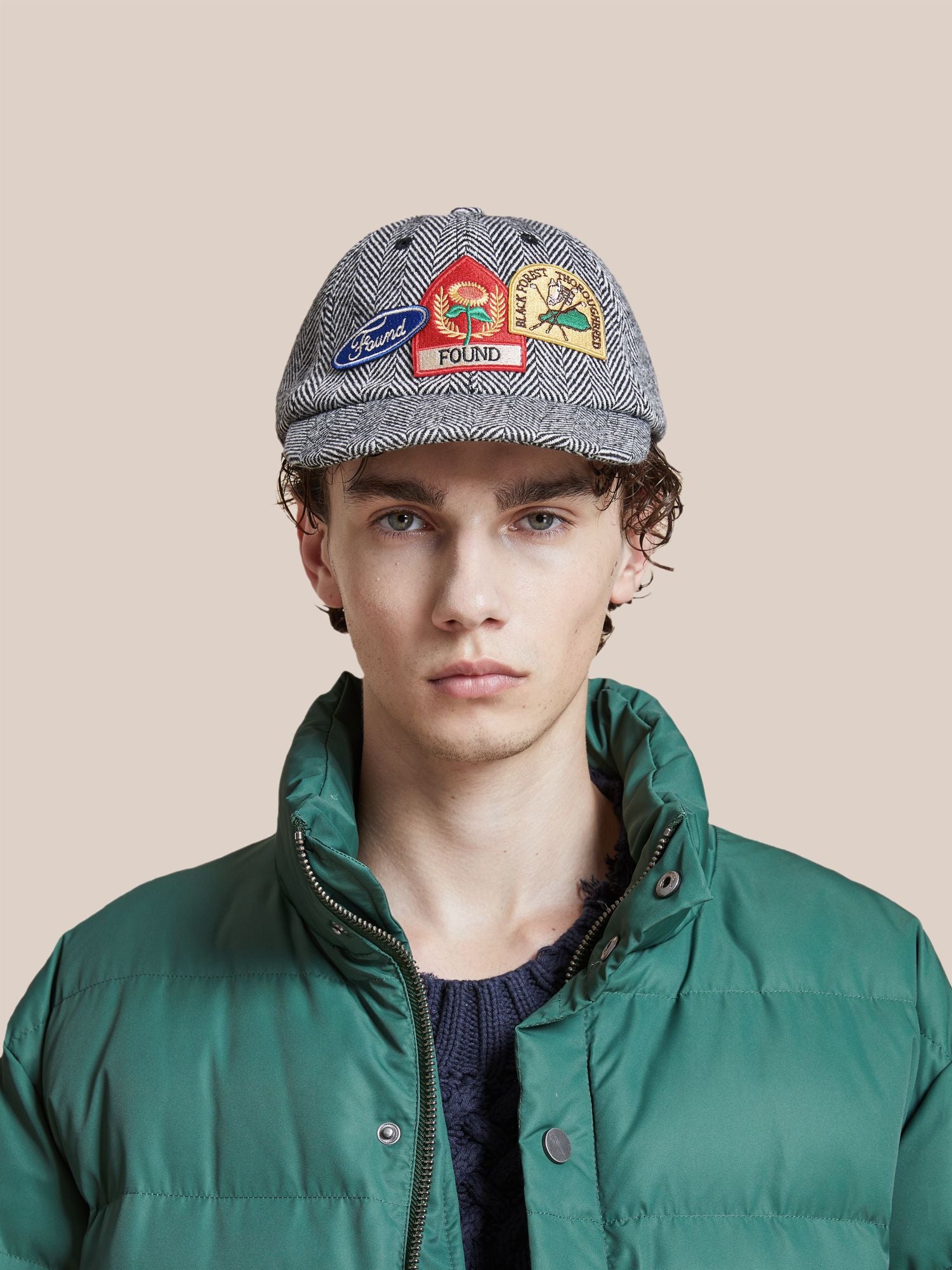 Young man with curly hair wearing a Found Herringbone Tweed Patch Cap and a green jacket, looking directly at the camera against a neutral background.