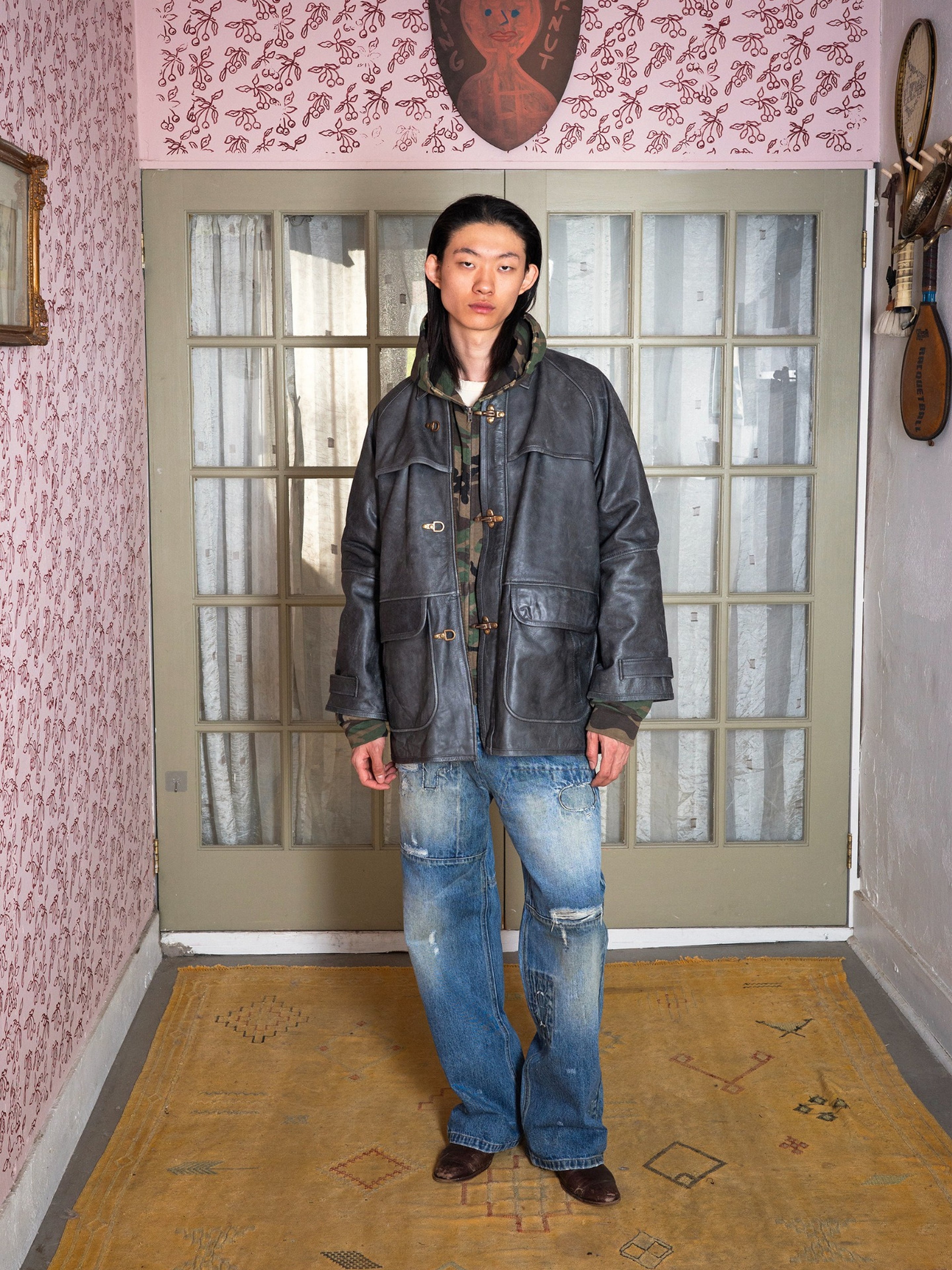A person indoors wears the FOUND Distressed Leather Fireman Clasp Jacket in a unisex relaxed fit, layered over clothing and paired with distressed blue jeans, set against glass doors and vintage decor.